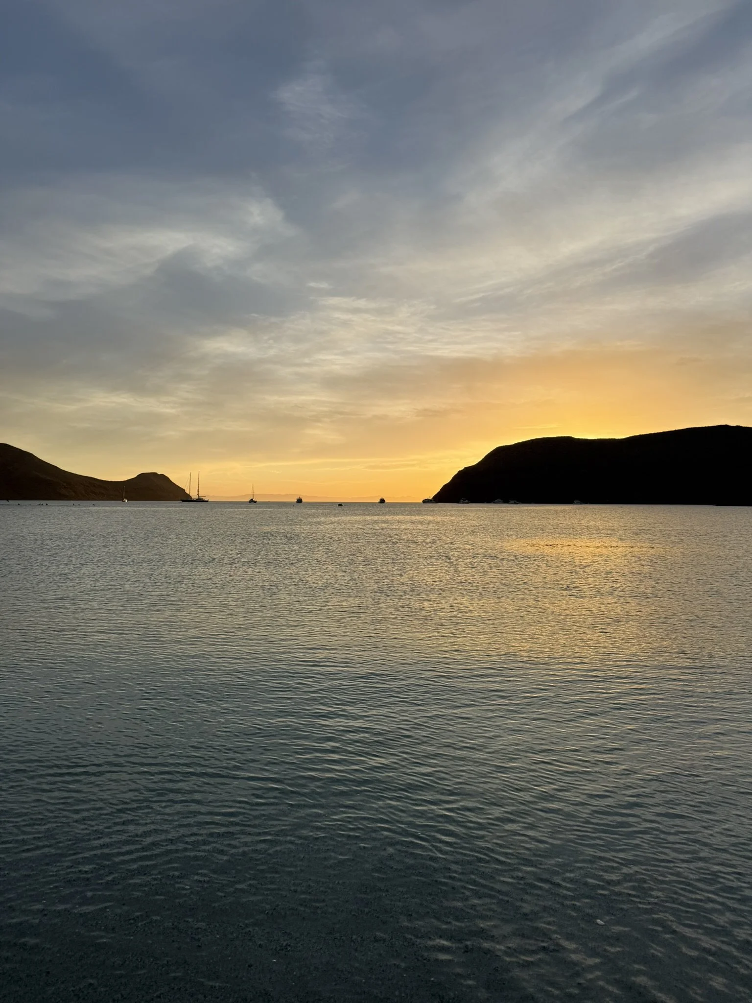 Sunset over calm water with distant hills and sailboats.
