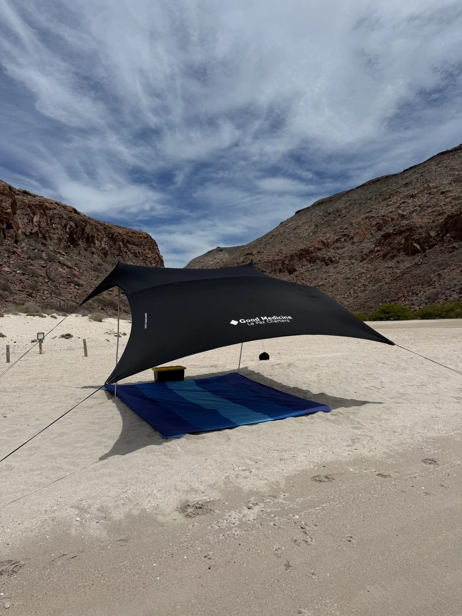 A desert landscape with a large umbrella shade structure and blanket on sandy ground, surrounded by rocky hills and a partly cloudy sky.