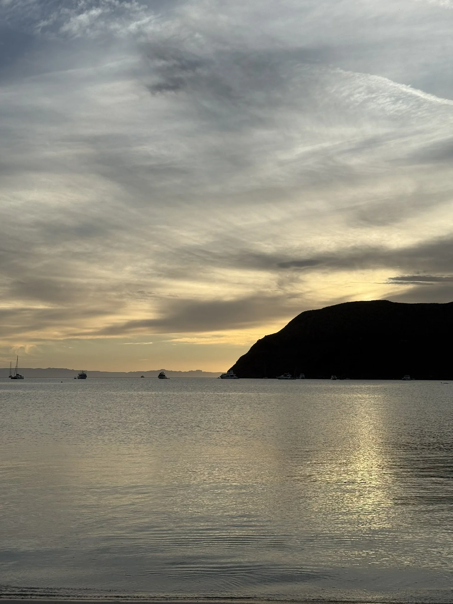 A serene body of water at sunset with a dark silhouette of a hill or mountain on the right, several boats anchored in the distance, and a cloudy sky with hints of orange and yellow near the horizon.