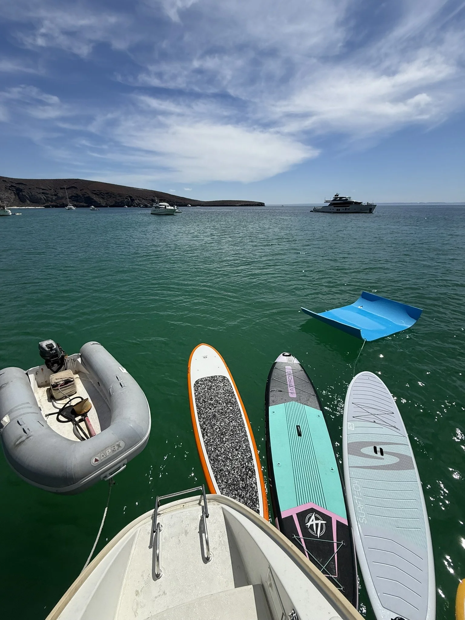 View of several paddleboards and boats floating on calm green water near a rocky shoreline on a partly cloudy day.