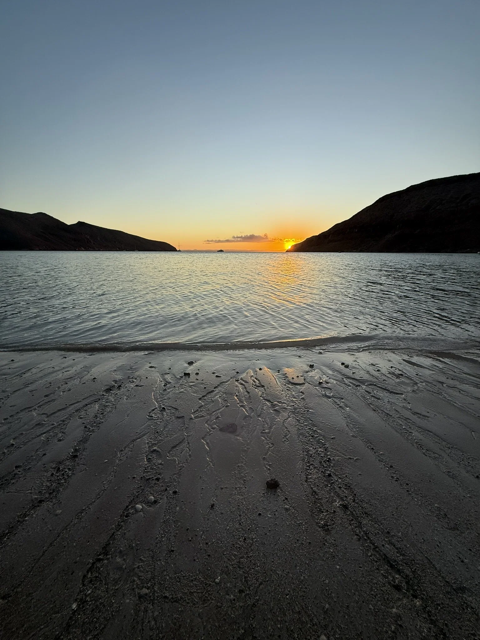 Sunset over a body of water with hills on both sides and a boat in the distance, sandy beach with scattered rocks in the foreground.