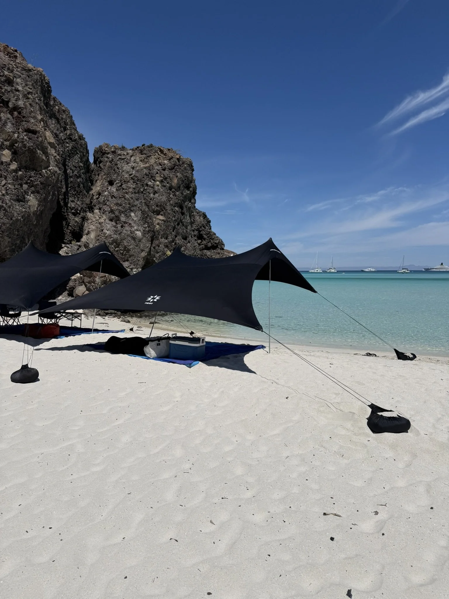 Beach scene with black umbrellas, rocky cliffs, turquoise water, and boats in the distance on a sunny day.