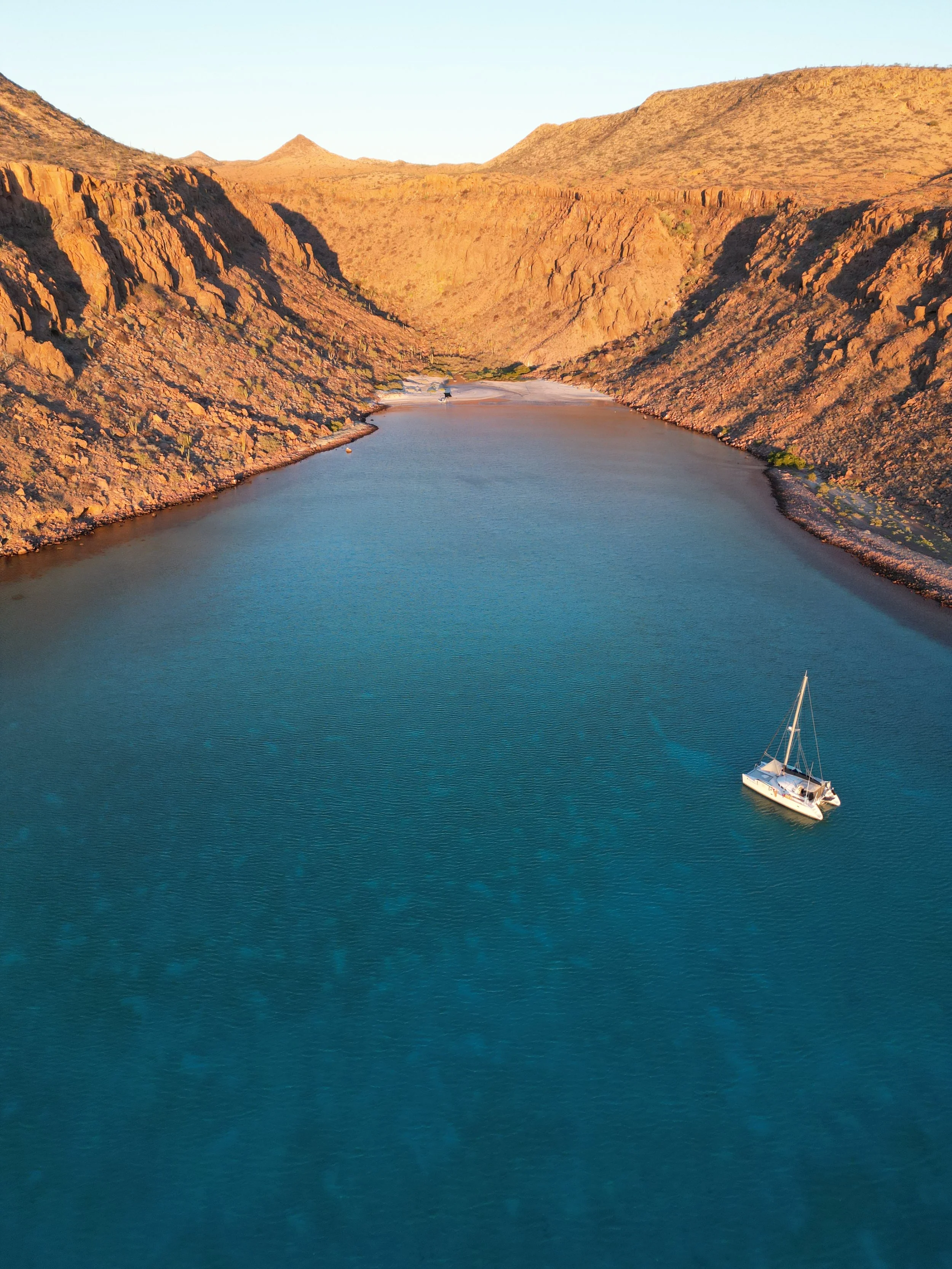 A sailboat on a calm body of water surrounded by rocky desert mountains at sunset.