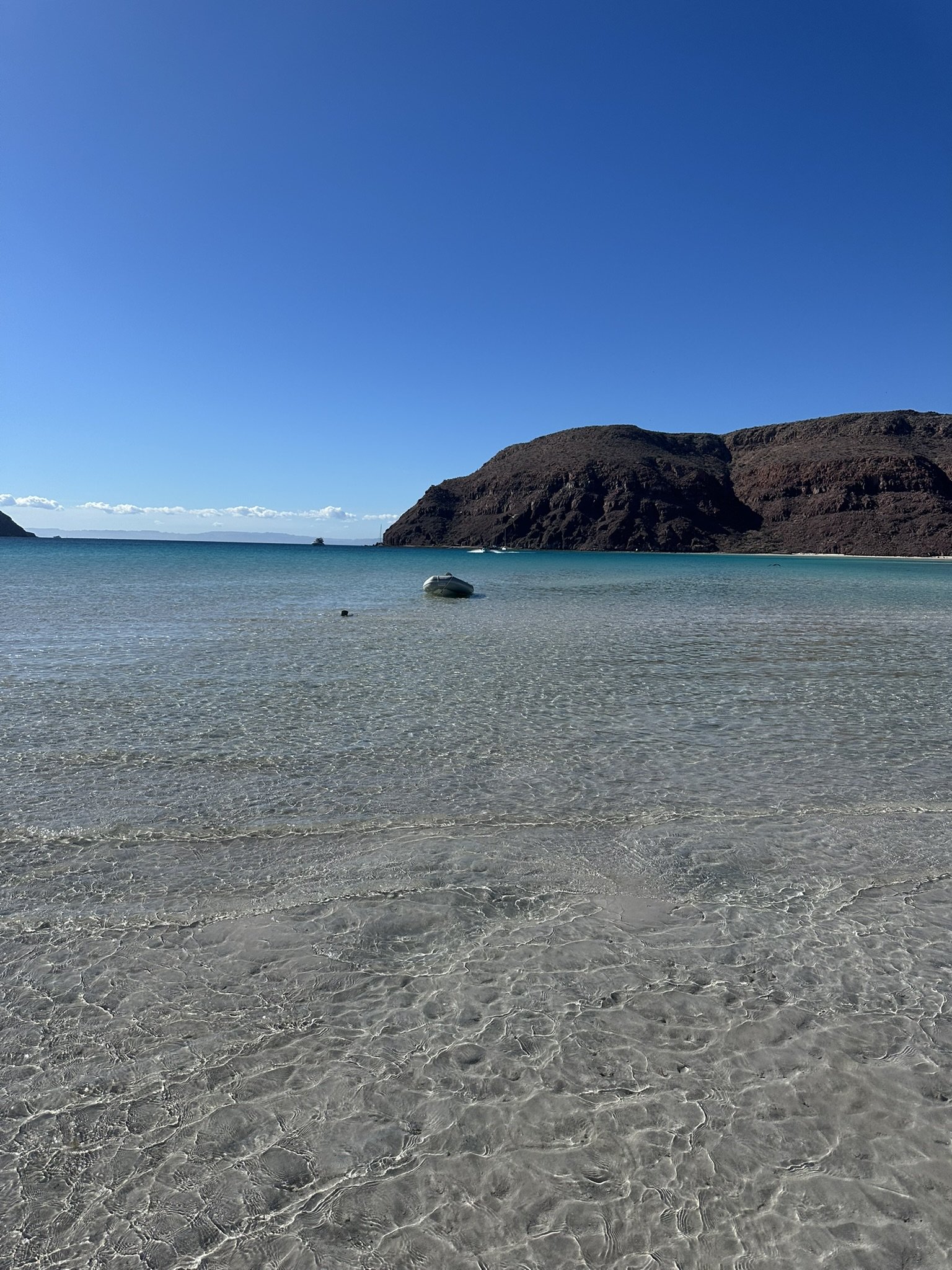 A clear, shallow beach with a small boat floating on the water, surrounded by rocky hills under a bright blue sky.
