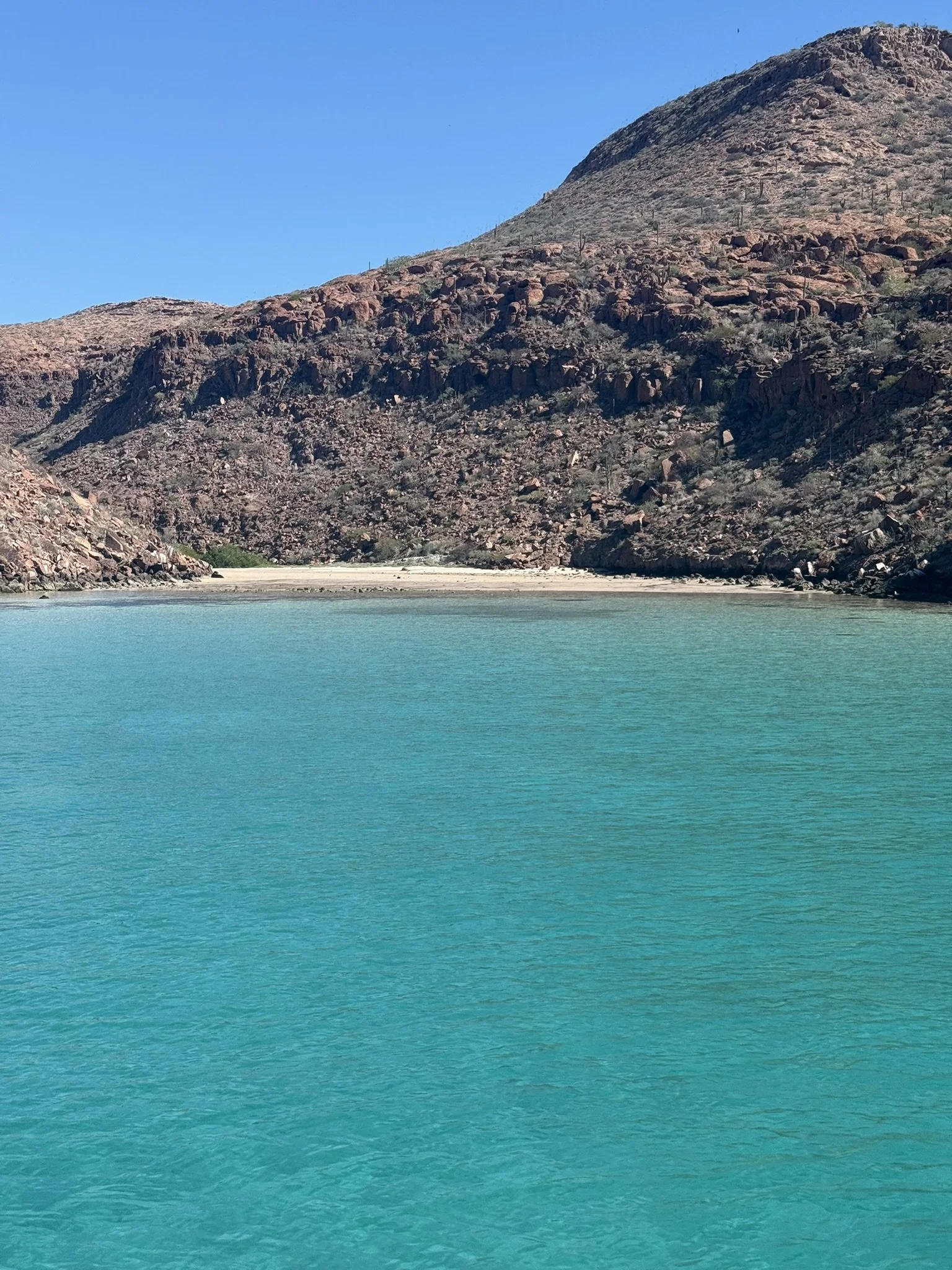Clear turquoise water in a lake with a mountain covered in rocks and sparse vegetation in the background against a blue sky.