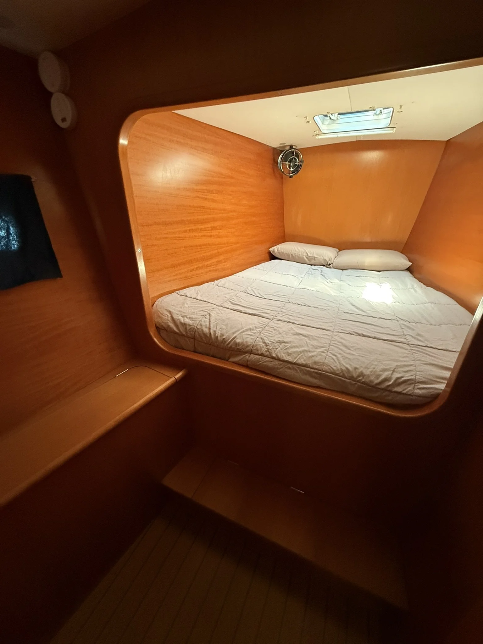 A cozy sleeping area on a boat or yacht with a wooden interior, a bed with white sheets and pillows, a small window, and a ceiling-mounted fan.
