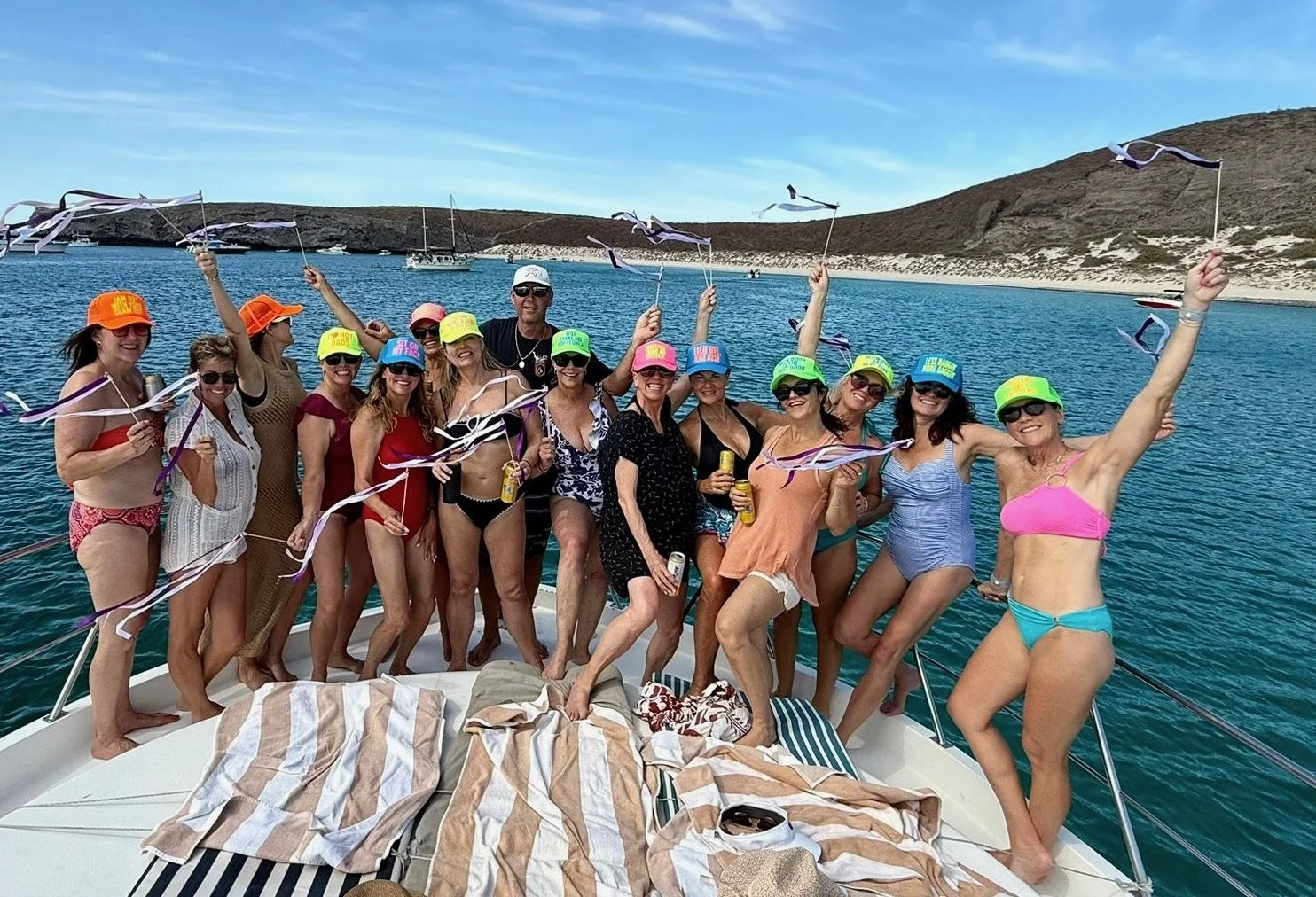 A group of women and one man celebrating on a boat, wearing swimsuits and colorful hats, holding drinks and waving ribbons, with a scenic ocean view and distant islands in the background.