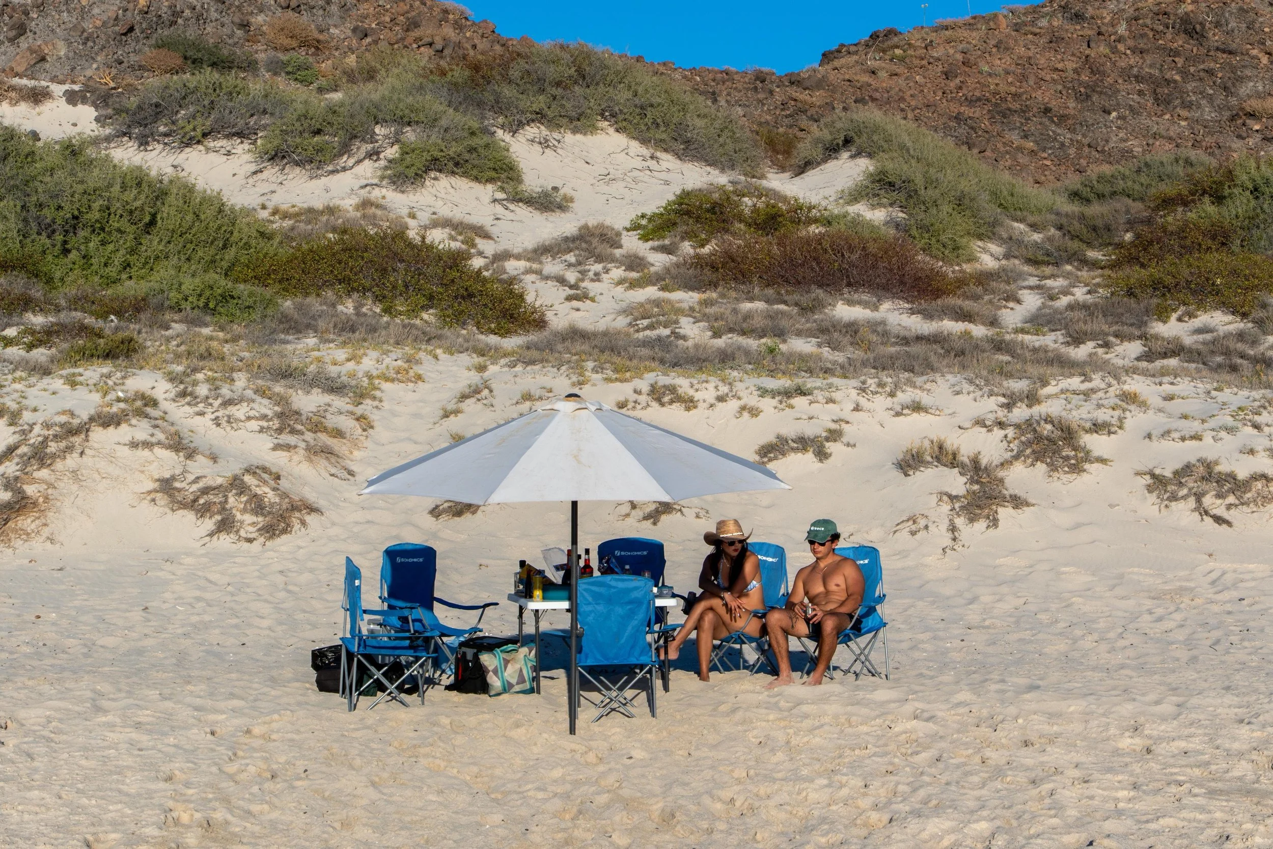 A woman and a man are sitting on camping chairs under a white beach umbrella on a sandy beach with desert shrubs and a hillside in the background.