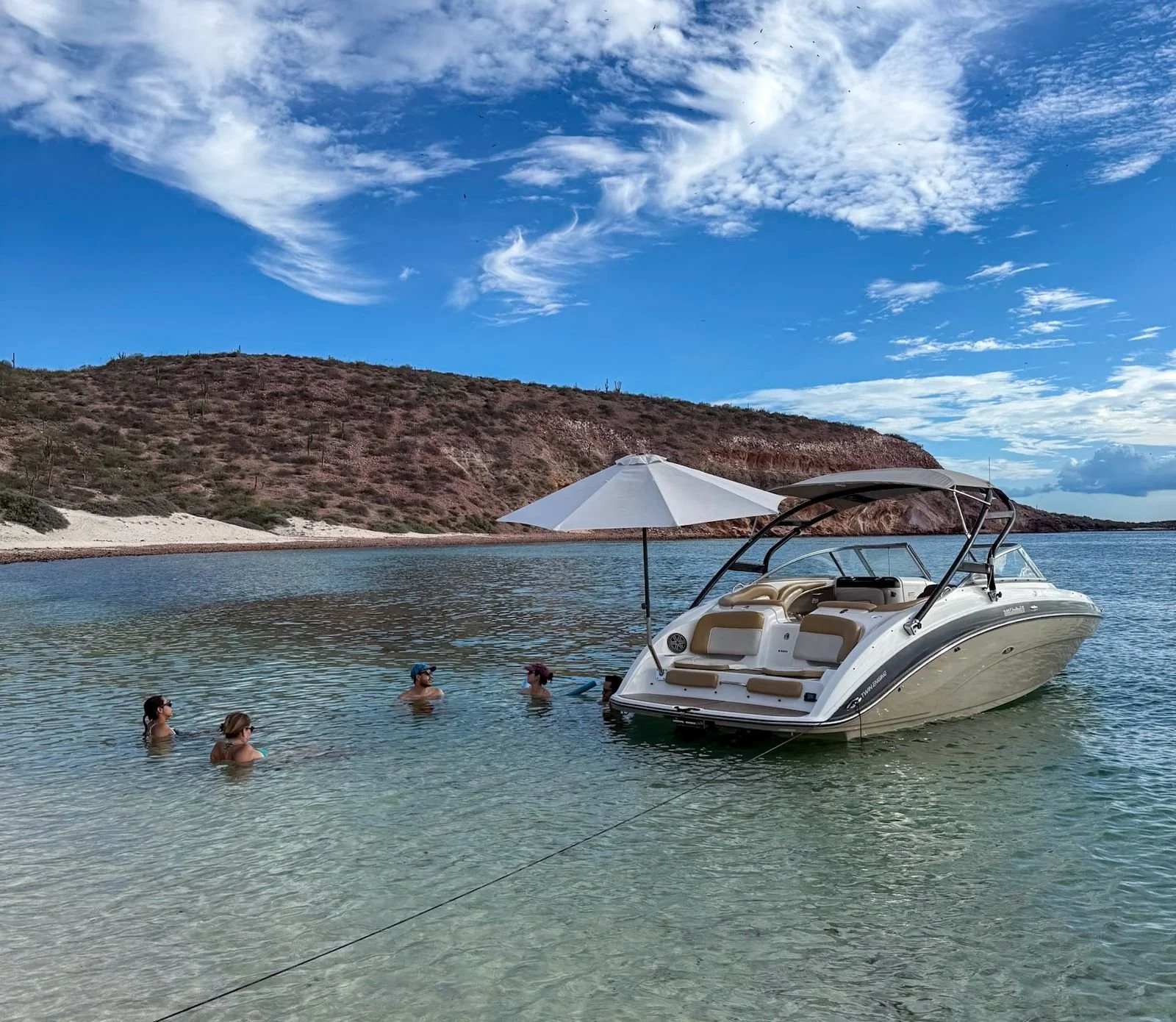 People swimming in a clear lake with a boat and an umbrella, surrounded by a hilly landscape and blue sky with clouds.