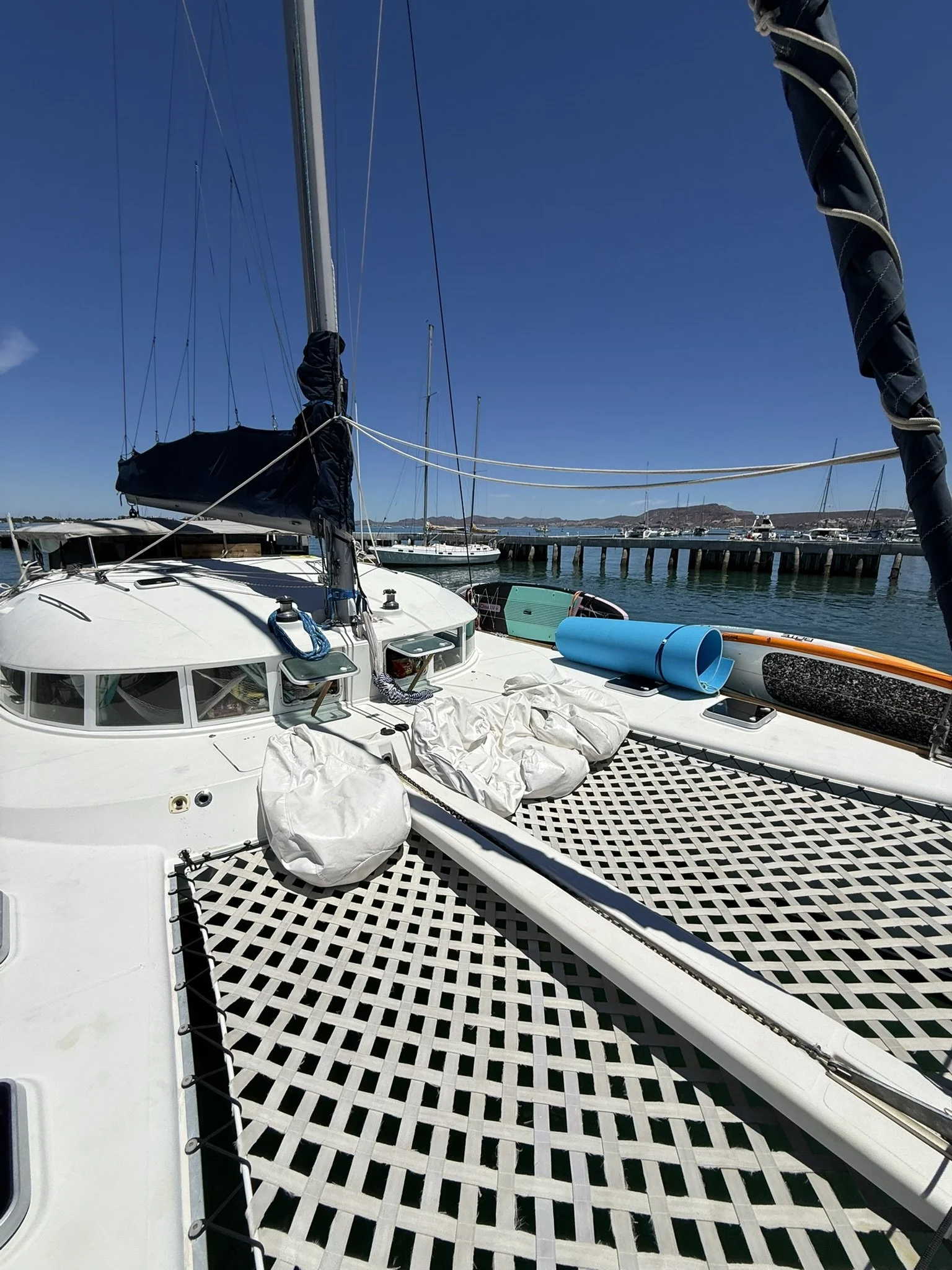 View of a docked sailboat with a white deck, black sail cover, blue water, and multiple boats in the background, under a clear blue sky.