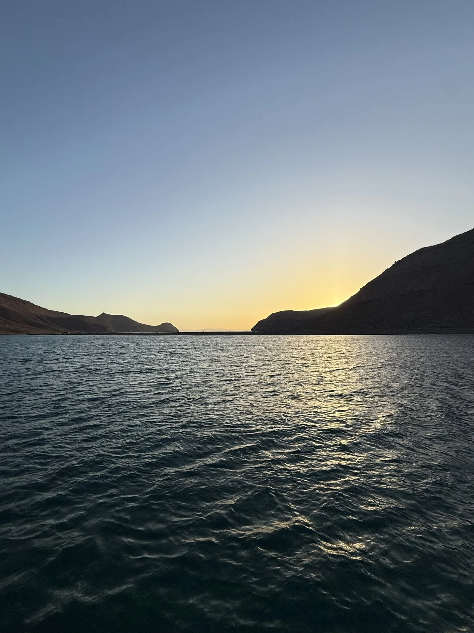 A body of water with ripples, surrounded by hills under a sky with the sun setting or rising.