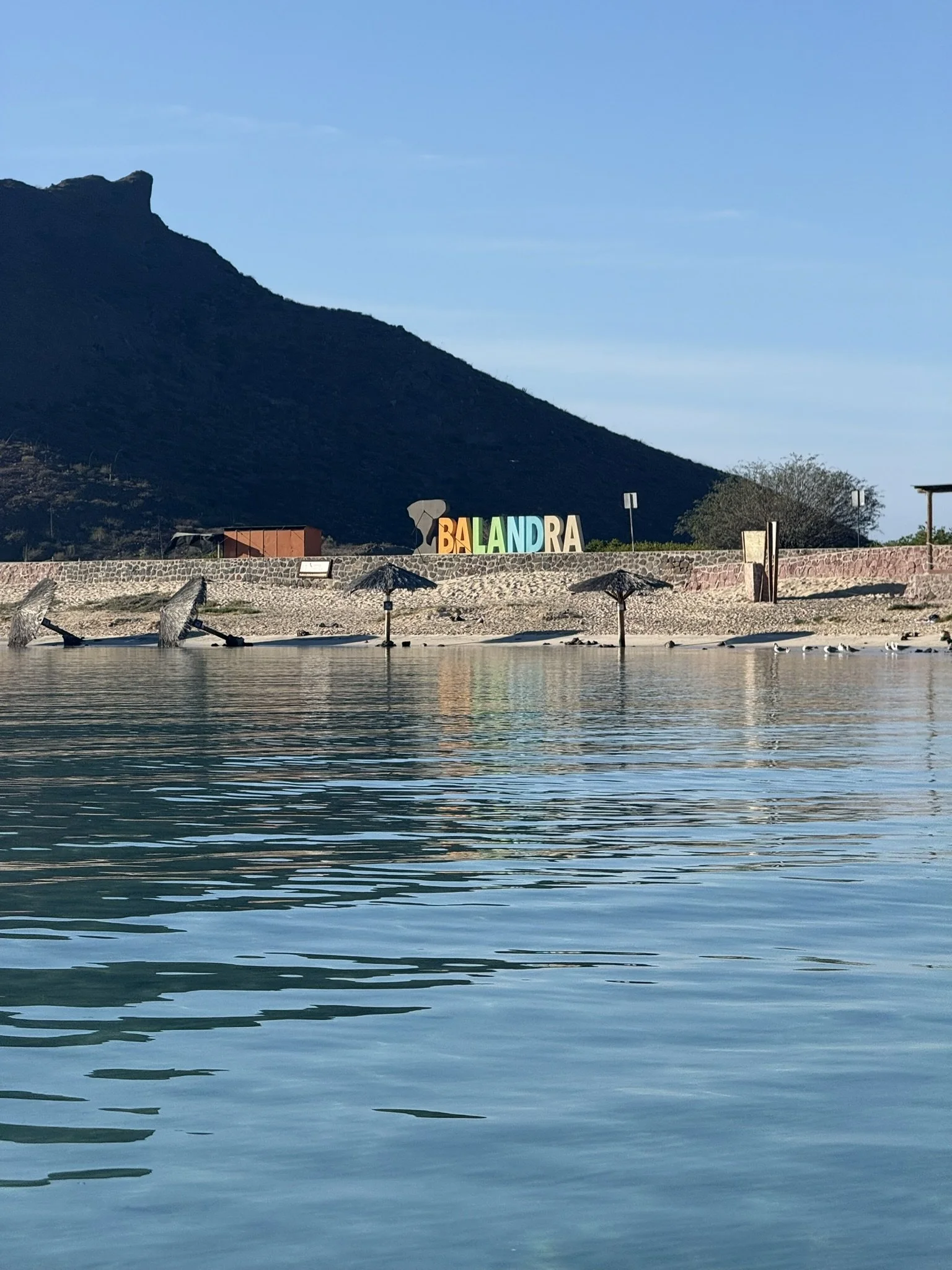 A scenic view of a body of water with gentle ripples, reflecting the blue sky above. In the background, a mountain rises with dark green vegetation and a rocky outcrop at its peak. Along the shore, there are a few small thatched umbrellas and seagull