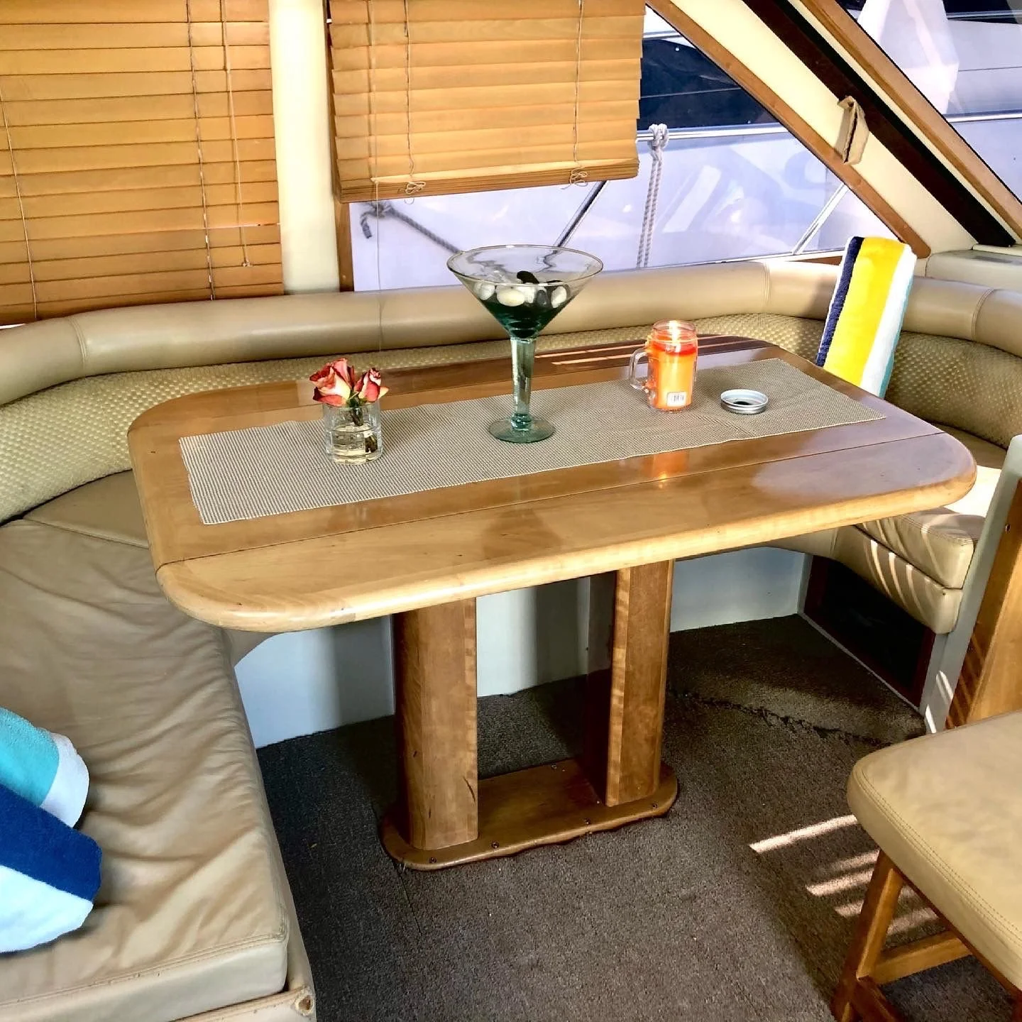 Boat interior with a corner dining area, wooden table, beige cushions, and decorative items including a martini glass, a small vase with roses, a candle, and a small container on the table.