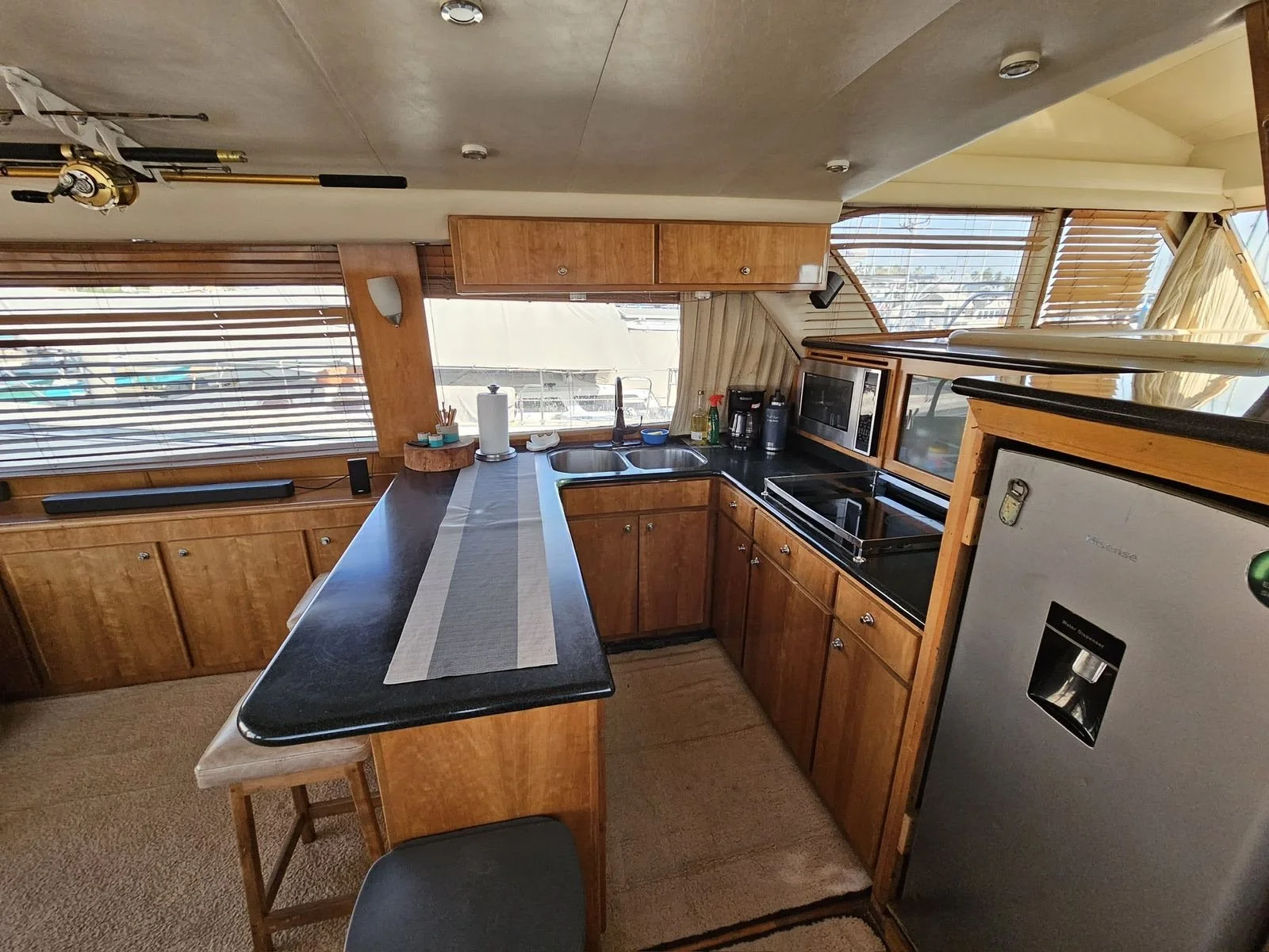 Kitchen with wooden cabinets, black countertop, microwave, refrigerator, double sink under large window, and some small appliances on the counter.