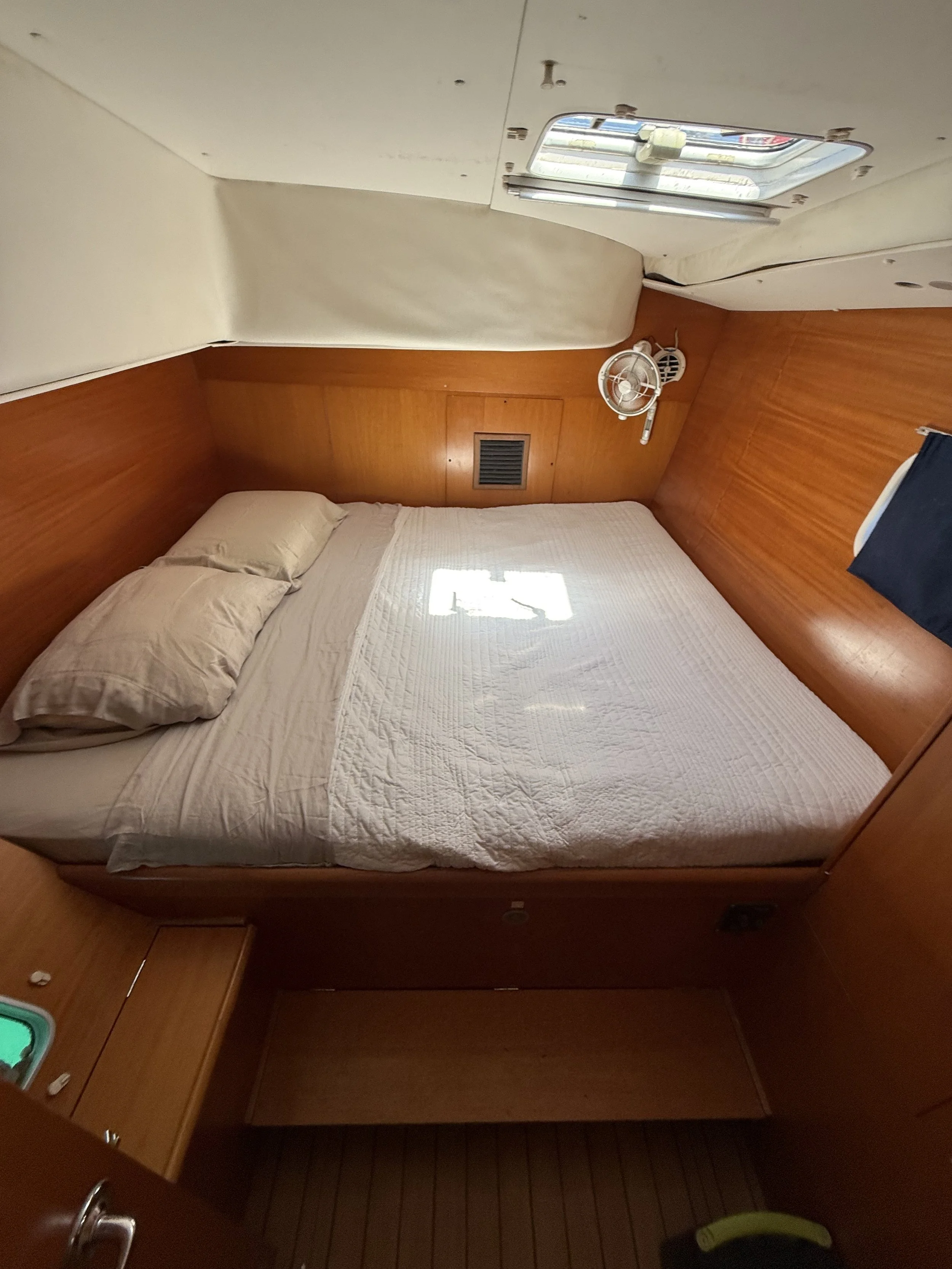 Inside a boat cabin showing a bed with beige pillows and a quilt, a wooden headboard, a small island with a light, a window, and a ceiling hatch.