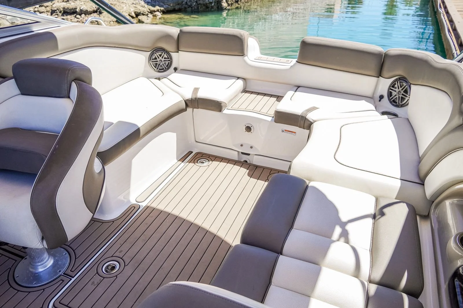 Luxury boat's seating area with beige and gray cushioned seats, built-in speakers, and a wooden deck, surrounded by clear water.