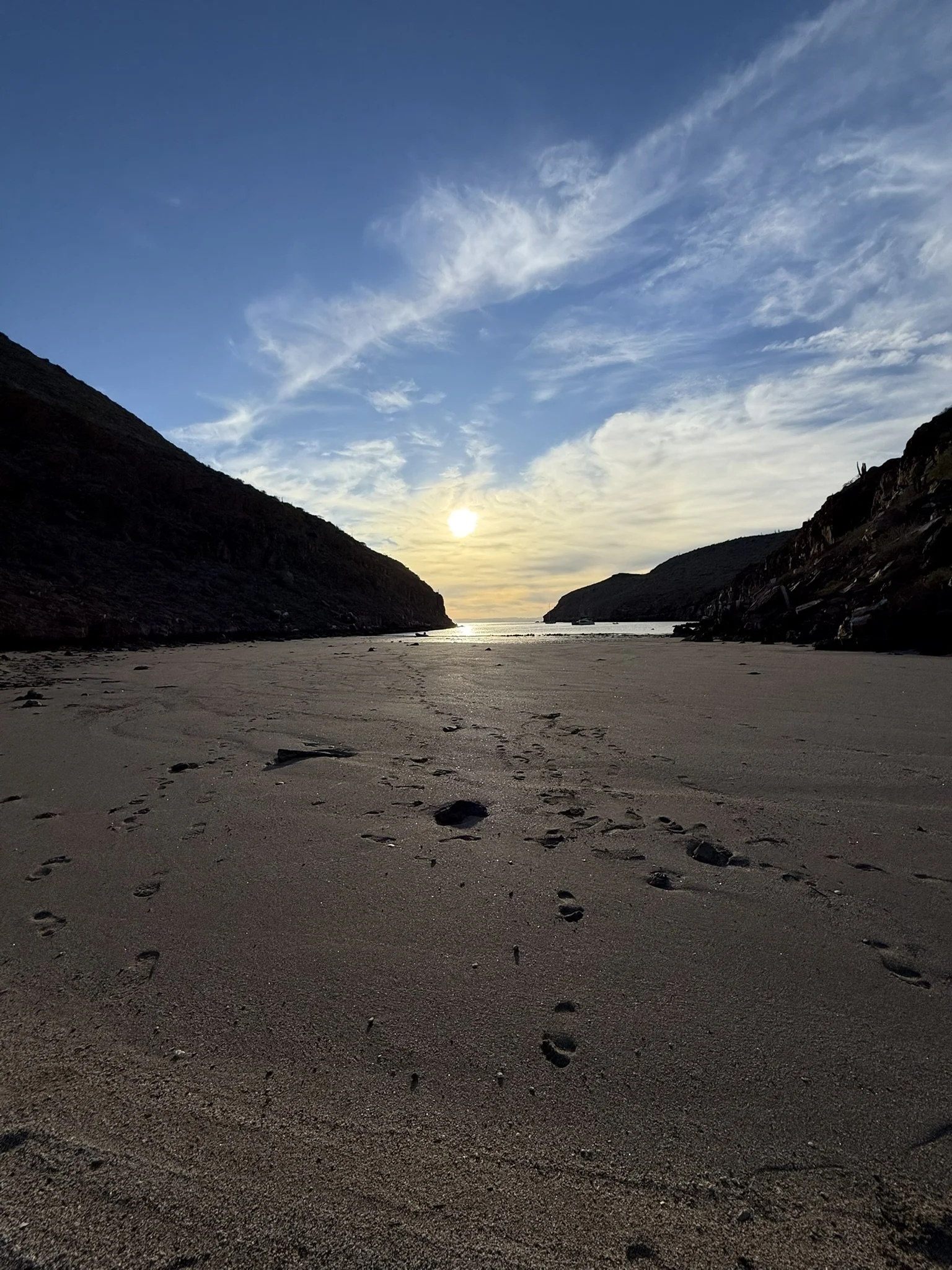 Sunset over a narrow river valley with rocky hills on each side and footprints in the sandy foreground.