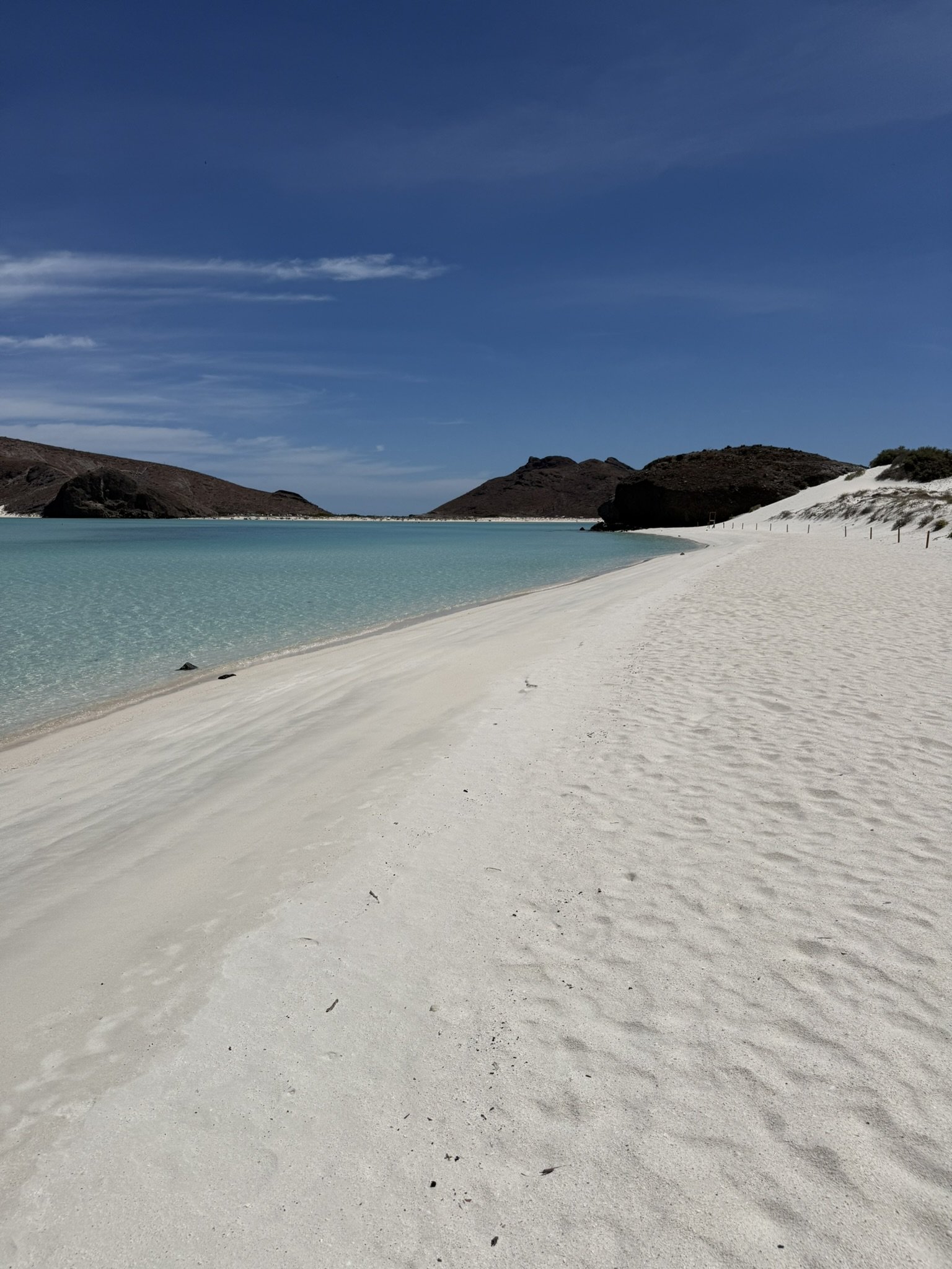 A serene beach with white sand, turquoise water, and distant brown hills under a partly cloudy blue sky.