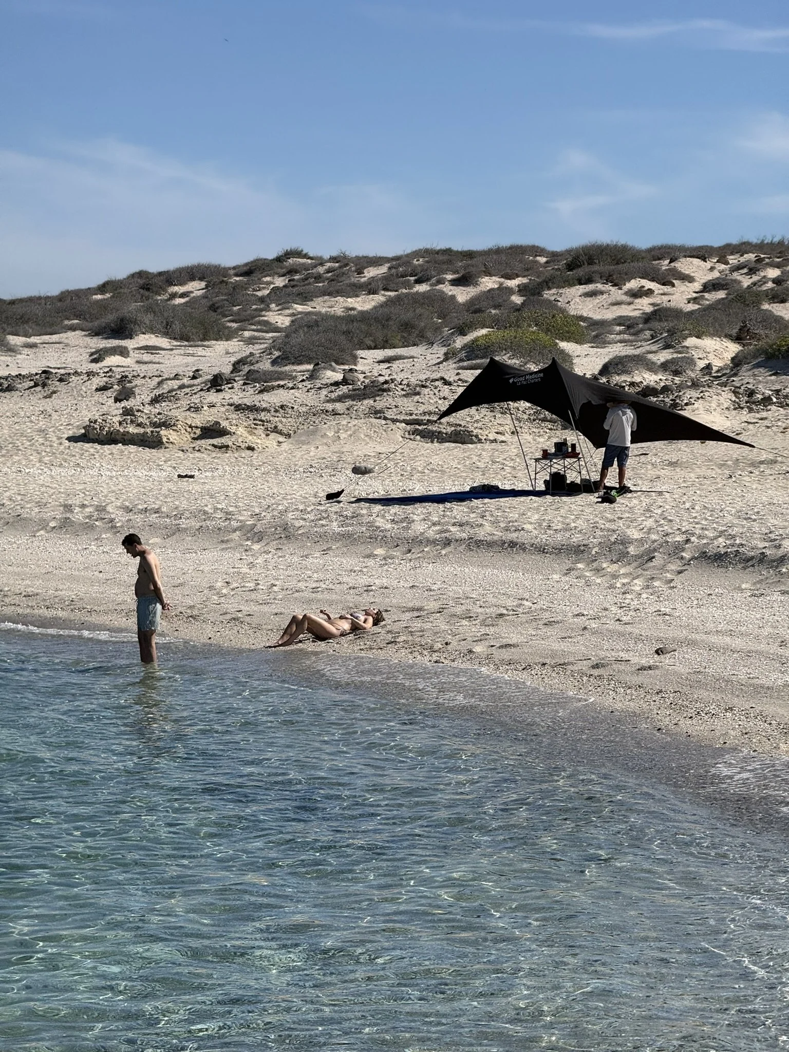 Beach scene with two people, one swimming in water and the other lying on the sand, with a man standing nearby. A shaded area is set up on the sand, and sand dunes with sparse vegetation are in the background.