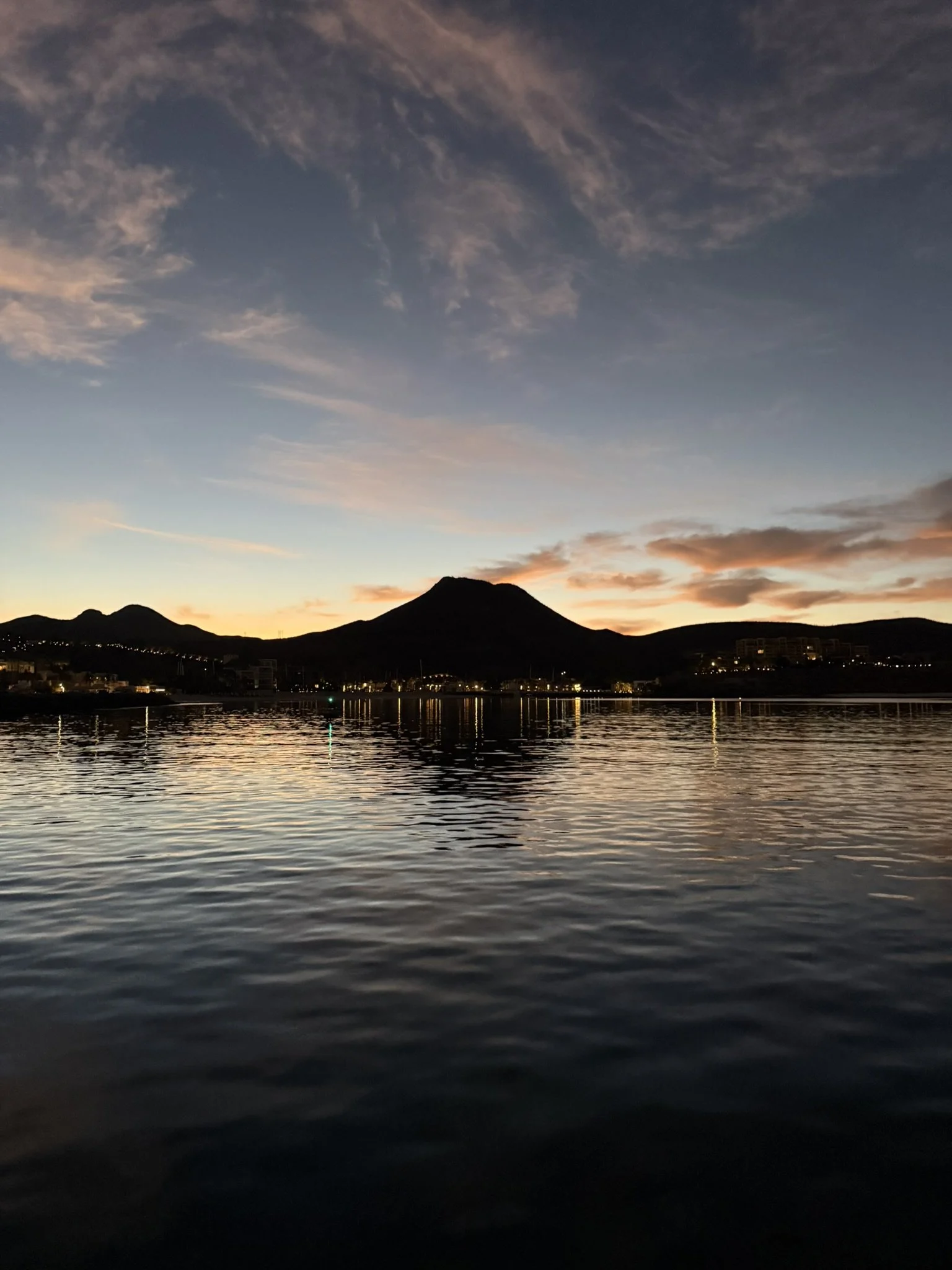 Sunset over a mountain and a body of water with small waves, reflection of city lights, and a colorful sky with clouds.
