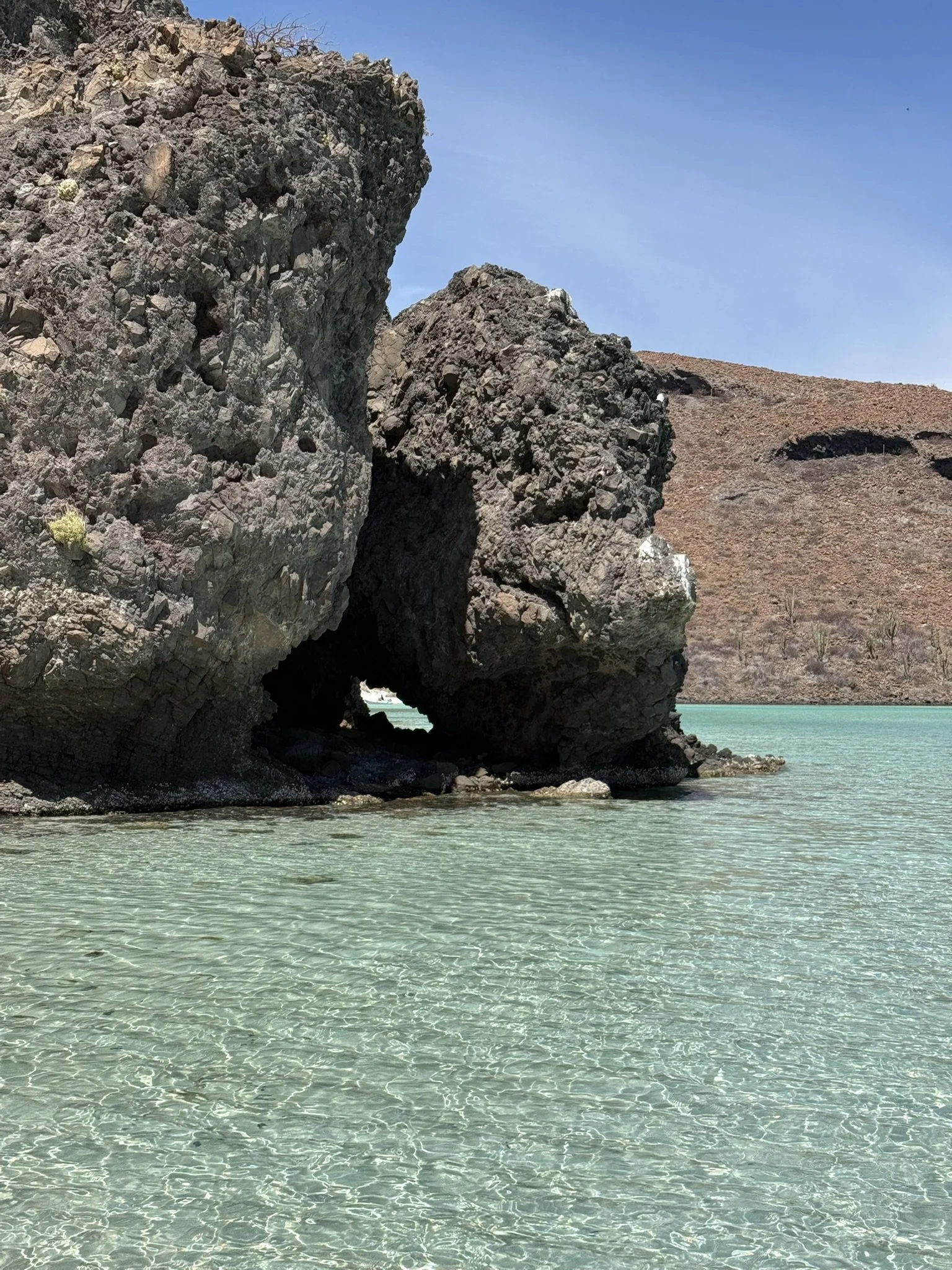 Large rocky formation with a natural archway near turquoise water, with a brown hillside and clear blue sky in the background.