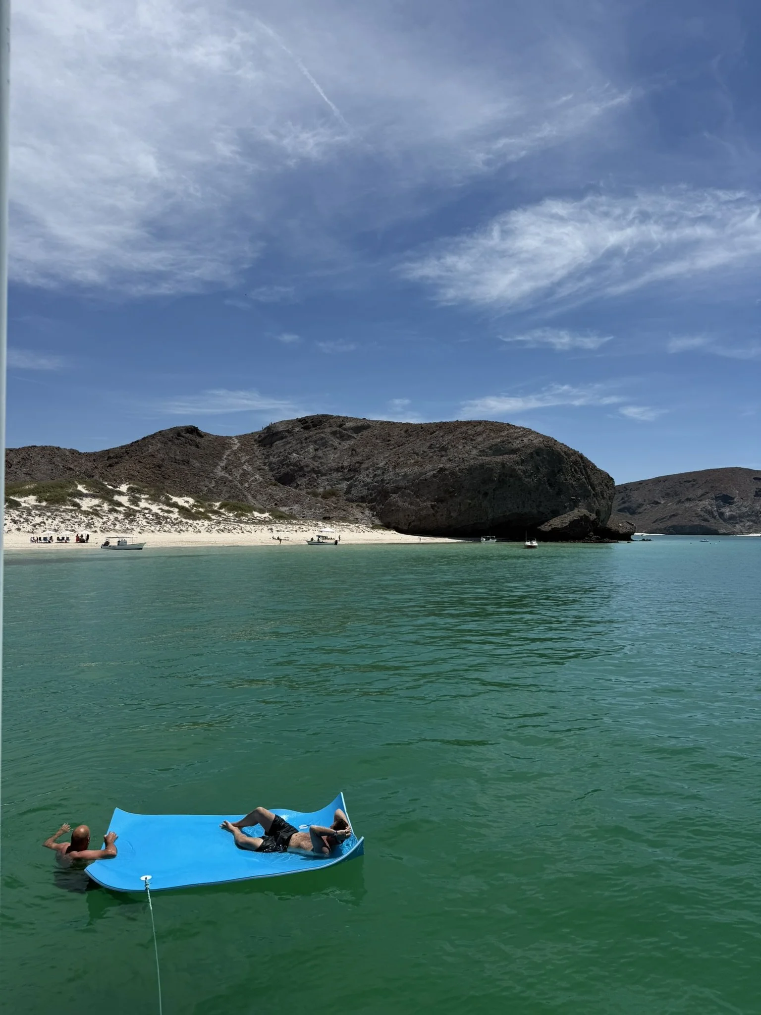 Two people relaxing on a blue float in a turquoise body of water near a sandy beach, with boats and rocky hills in the background under a partly cloudy sky.