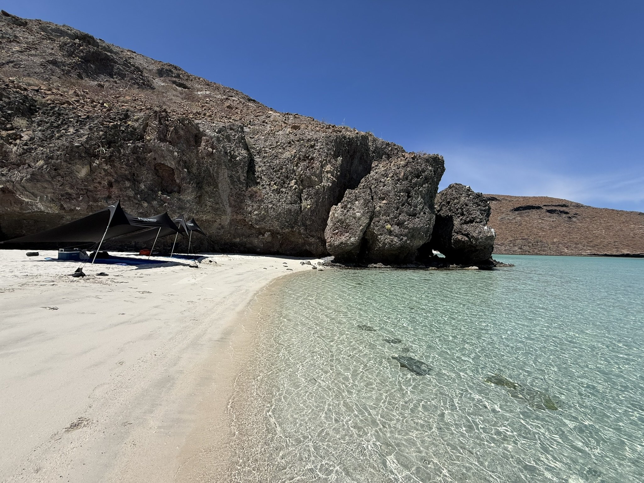 A tranquil beach with white sand, clear turquoise water, large rocks, and a canopy with shaded seating, against a backdrop of dry hills and a bright blue sky.