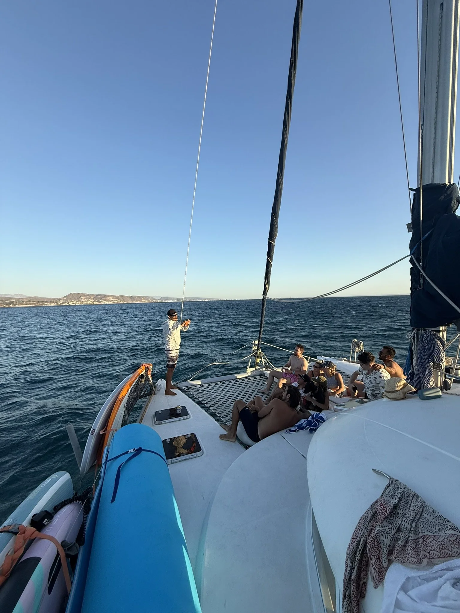People relaxing and enjoying a day on a sailboat, with a body of water and distant land visible in the background.