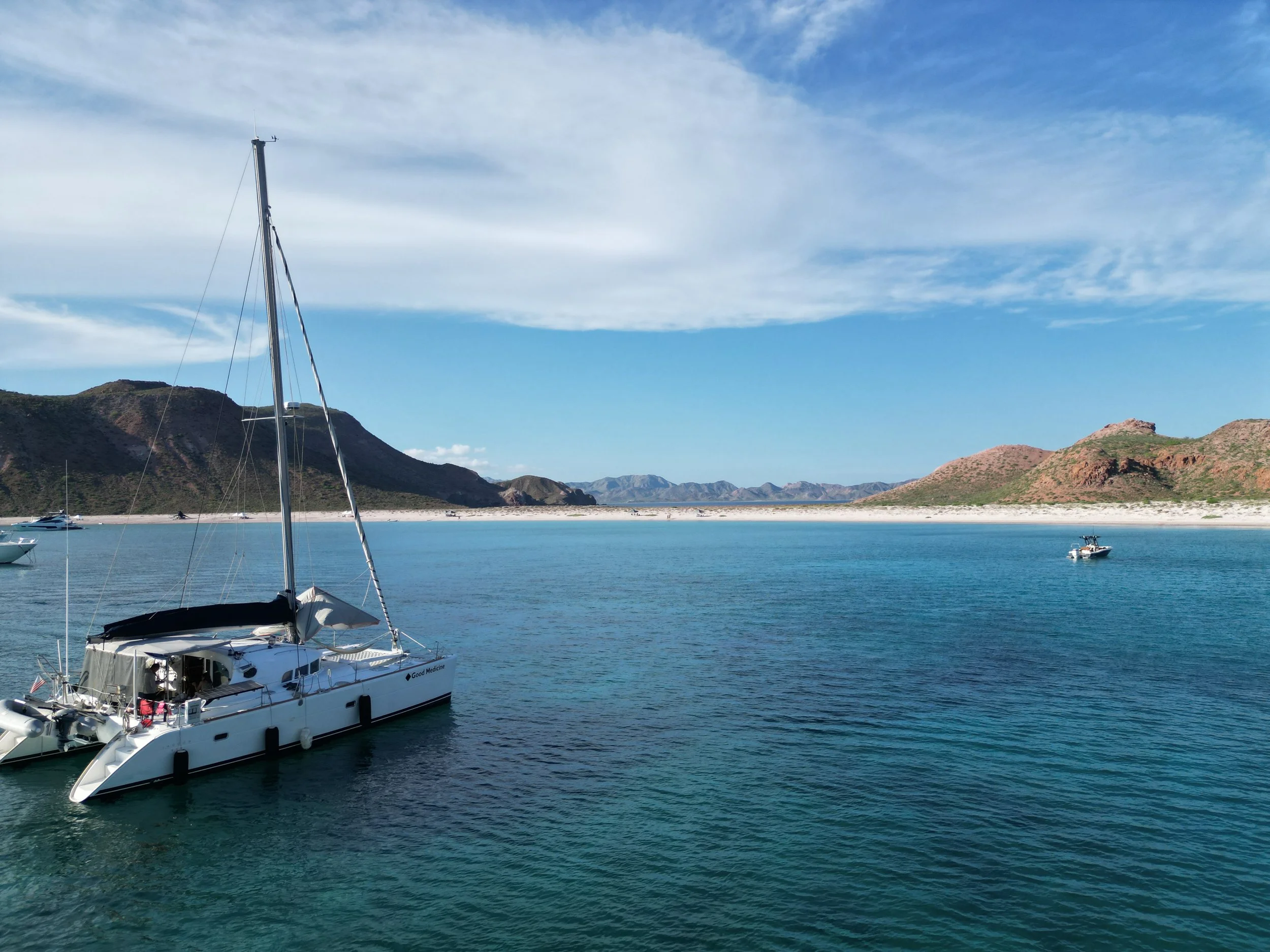 A large sailboat with a tall mast in a marina or harbor with clear blue water, mountains in the background, and a partly cloudy sky.