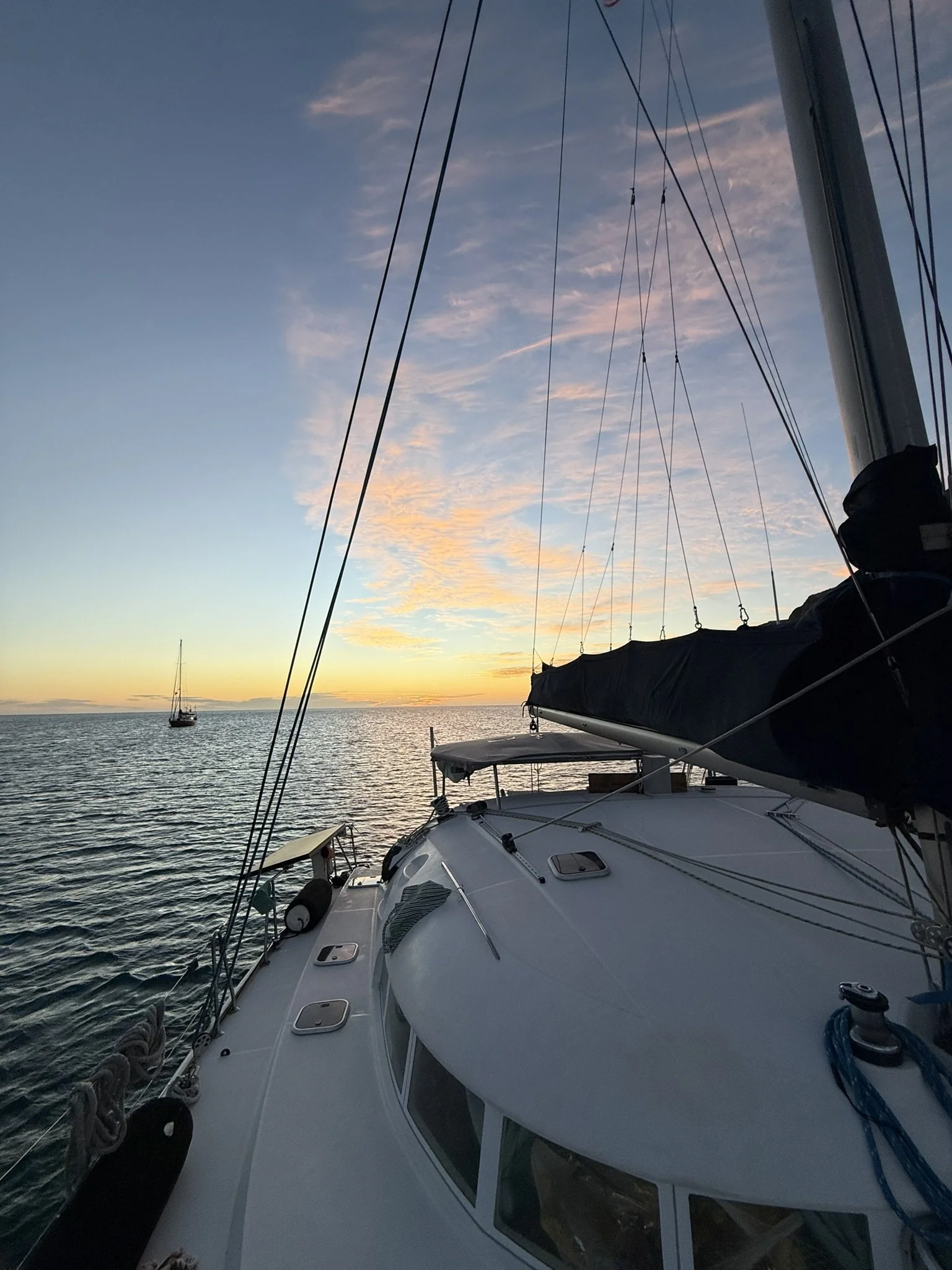 A sailboat on calm water during sunset with visible rigging and a covered sail, and another sailboat in the distance near the horizon.