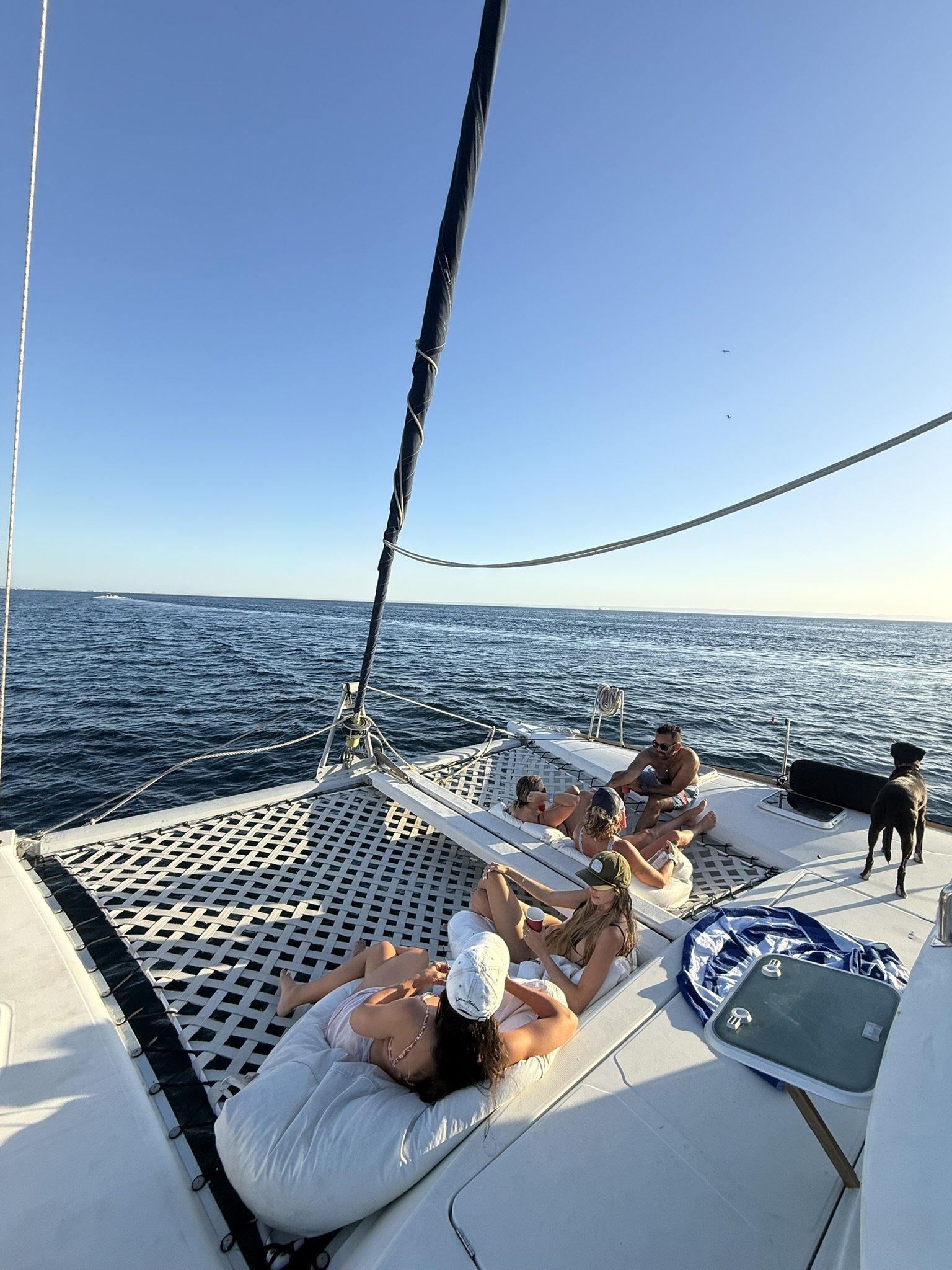 People relaxing and socializing on the deck of a boat during sunset, with some wearing hats and sunglasses, and the sea visible in the background.