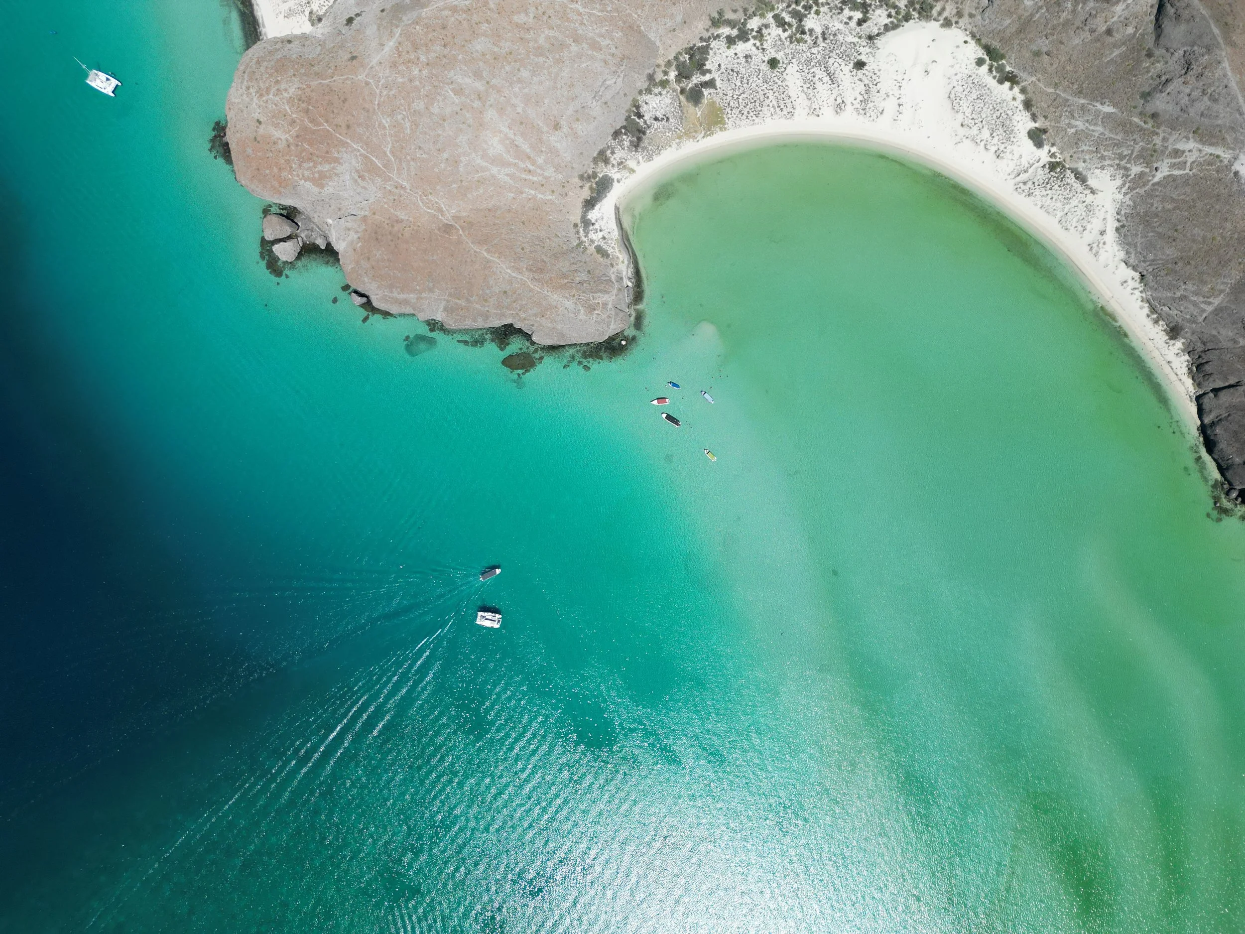 Aerial view of a coastal area with turquoise water, a white sandy beach, rocky cliffs, and boats in the water.