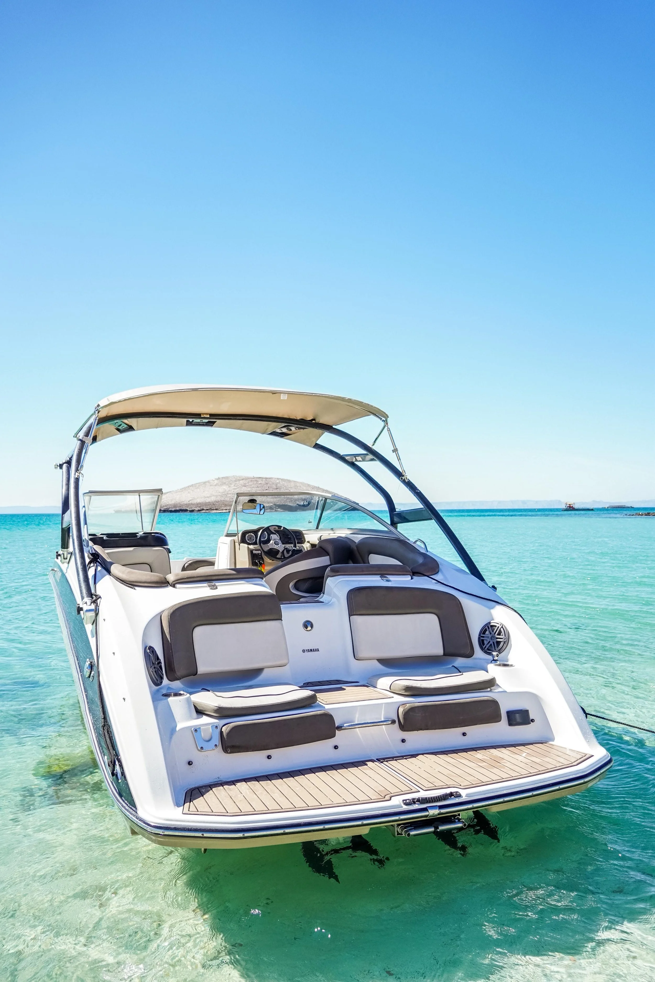 A white boat with black and beige seating floats on clear turquoise water near a sandy beach, with a small island and distant pier in the background under a blue sky.