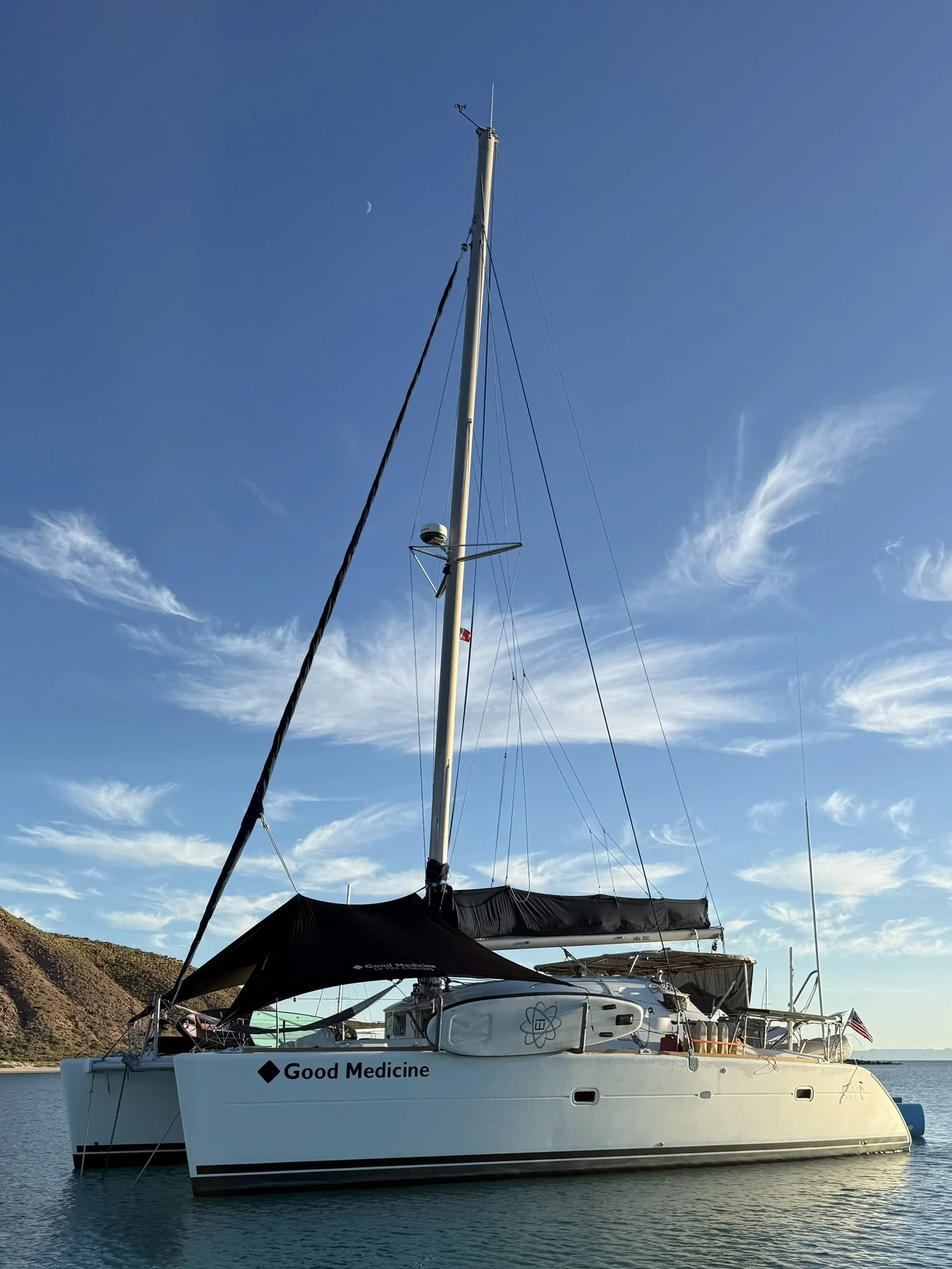 White sailing yacht with black canopy on calm water, with hills and a clear sky with some clouds and a crescent moon in the background.