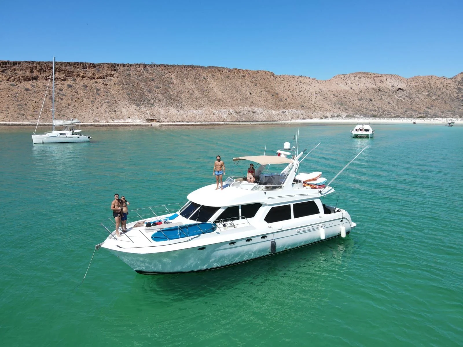 People enjoying a boat trip on a white yacht anchored in turquoise water with sailboats and a barren hillside in the background.