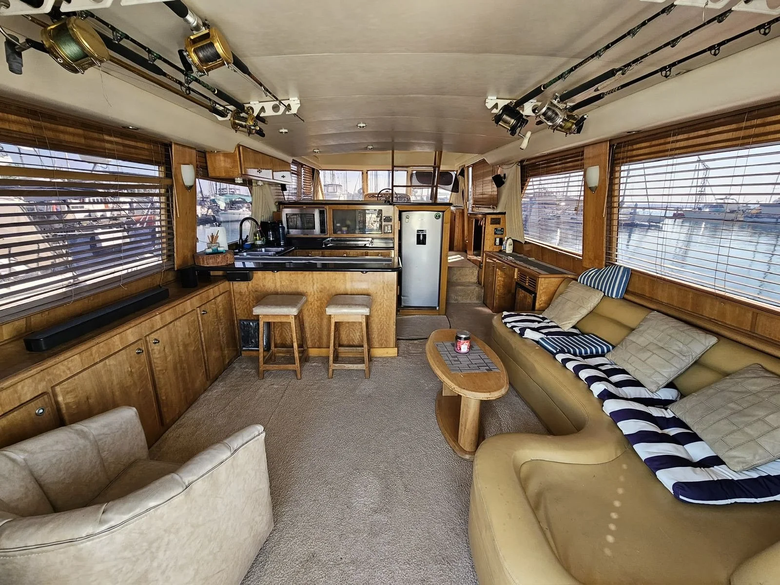 Interior of a boat with wooden paneling, a beige leather sofa with pillows, a small coffee table, and a kitchenette with stools, surrounded by large windows.