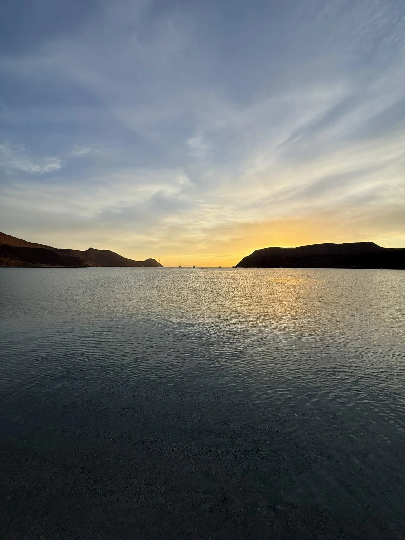 A peaceful body of water at sunset with hills on the horizon and a sailboat in the distance, capturing a serene natural landscape.