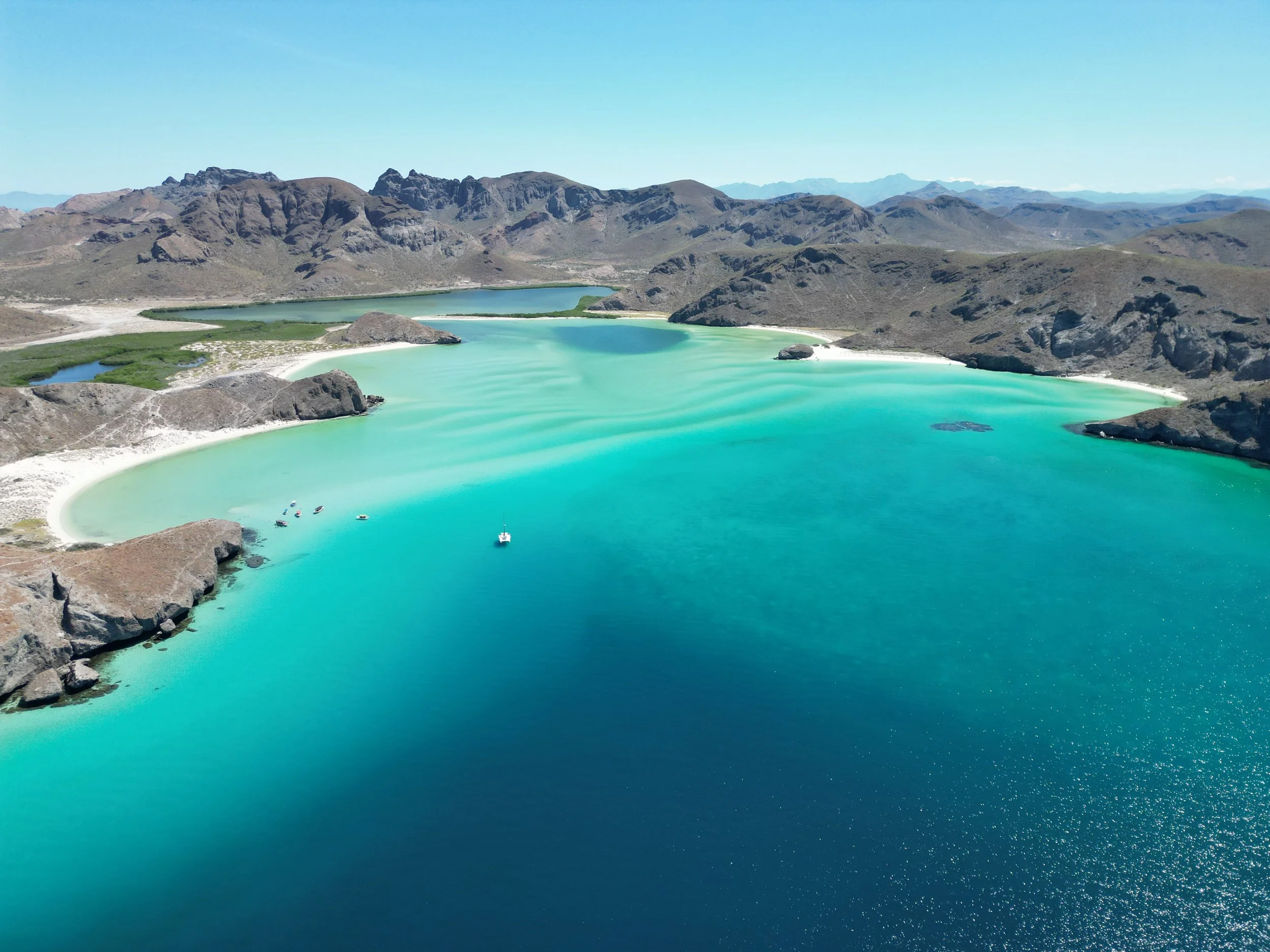 Aerial view of a turquoise lake with sandy beaches, surrounded by rugged brown mountains under a clear blue sky.
