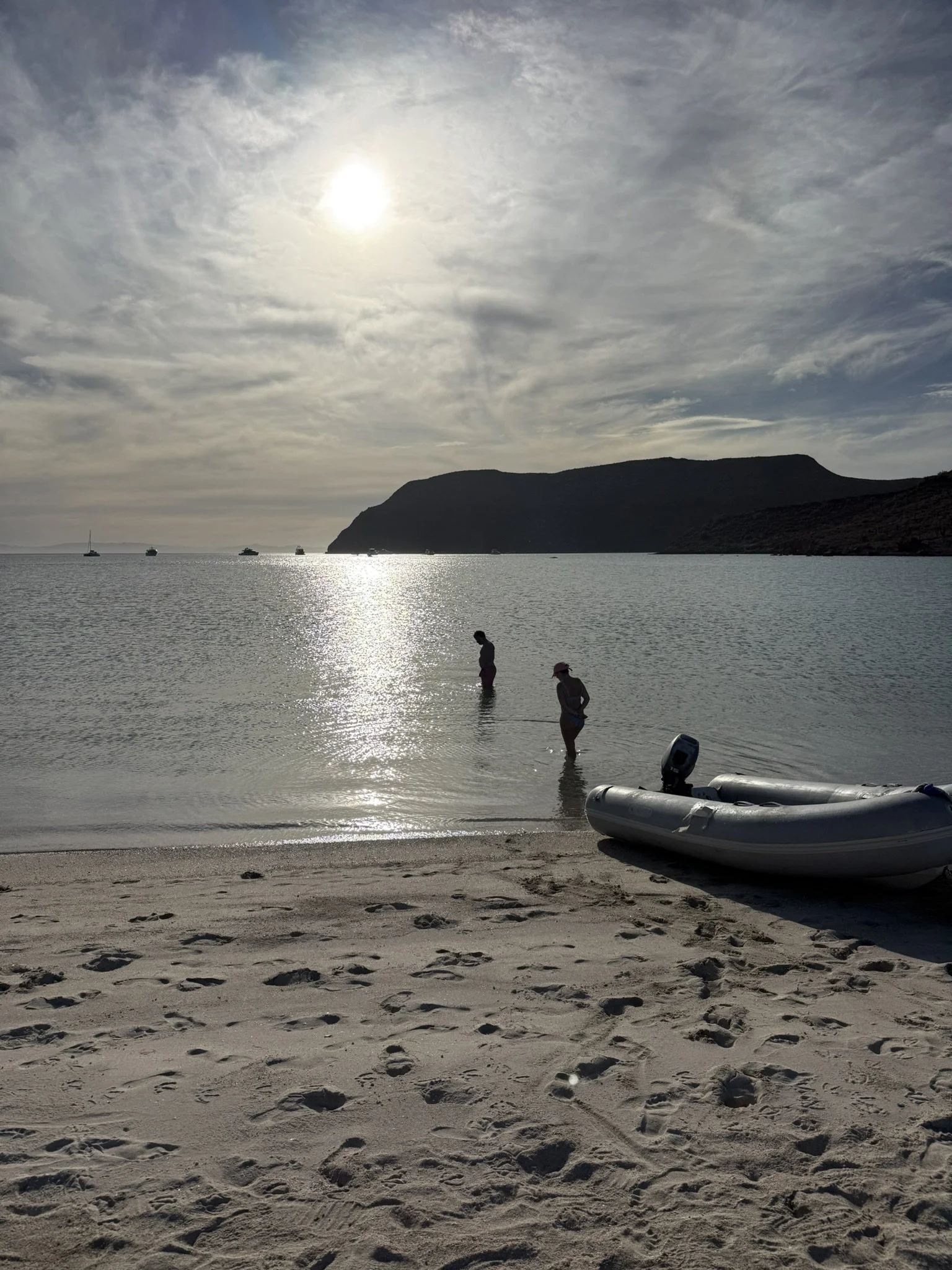 A beach scene during sunset with two children standing in shallow water, a small inflatable boat on the shoreline, and sailboats with a mountain in the background.