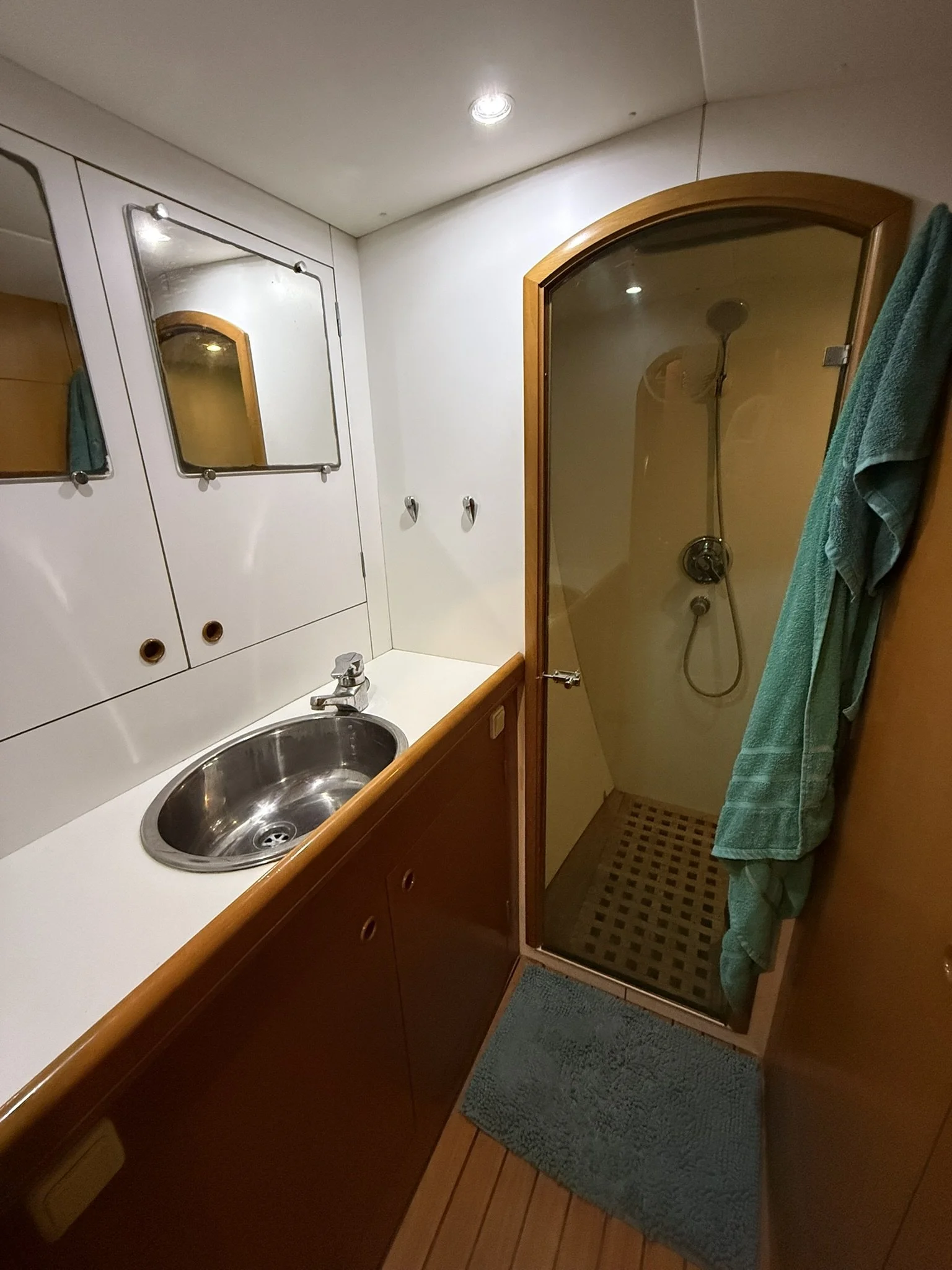 A compact bathroom with a small vanity sink, a mirror, and a shower enclosed by a wooden frame with a glass door. A teal towel hangs on a hook outside the shower, with a small green bath mat on the wooden floor.