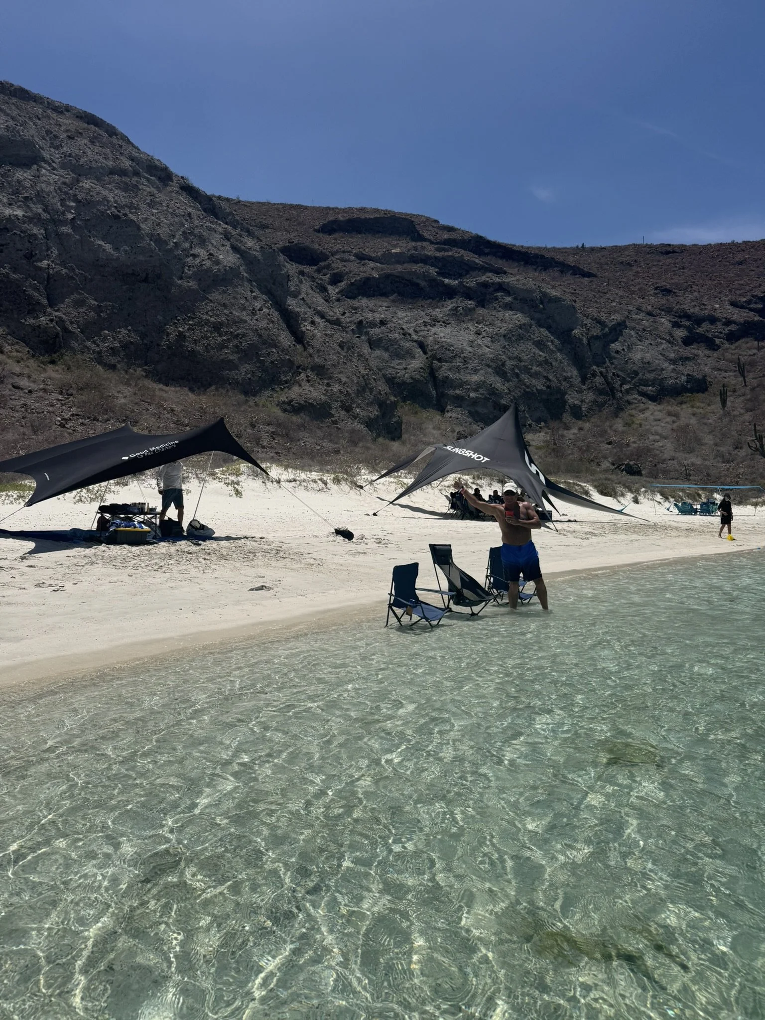 Beach scene with clear water, people under shade tents, rocky hills in background, and a man near chairs in the water.