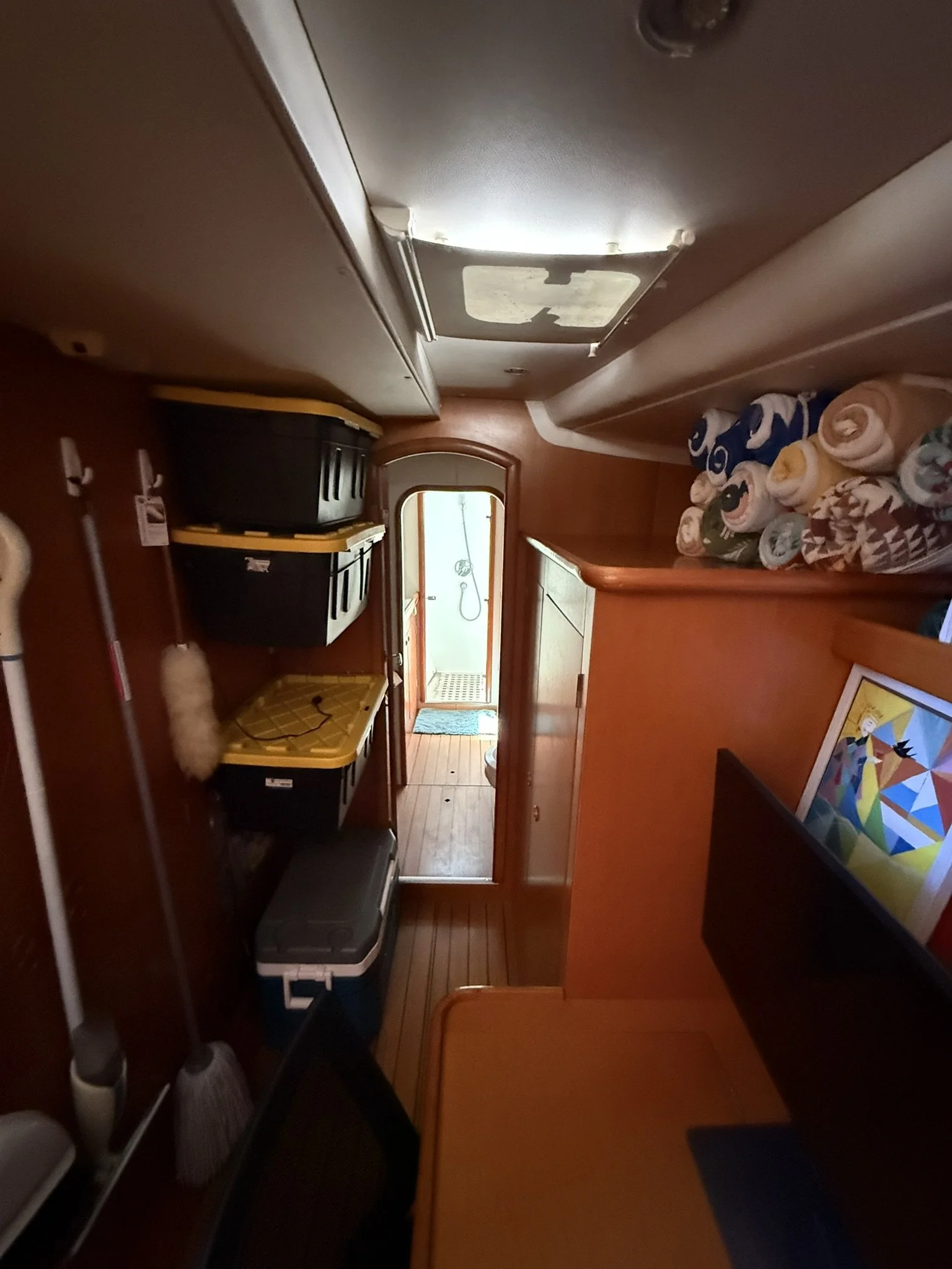 Interior view of a narrow hallway inside a boat or RV, with a small shower area at the end, rolled towels on a shelf to the right, and luggage and cleaning supplies on the left.