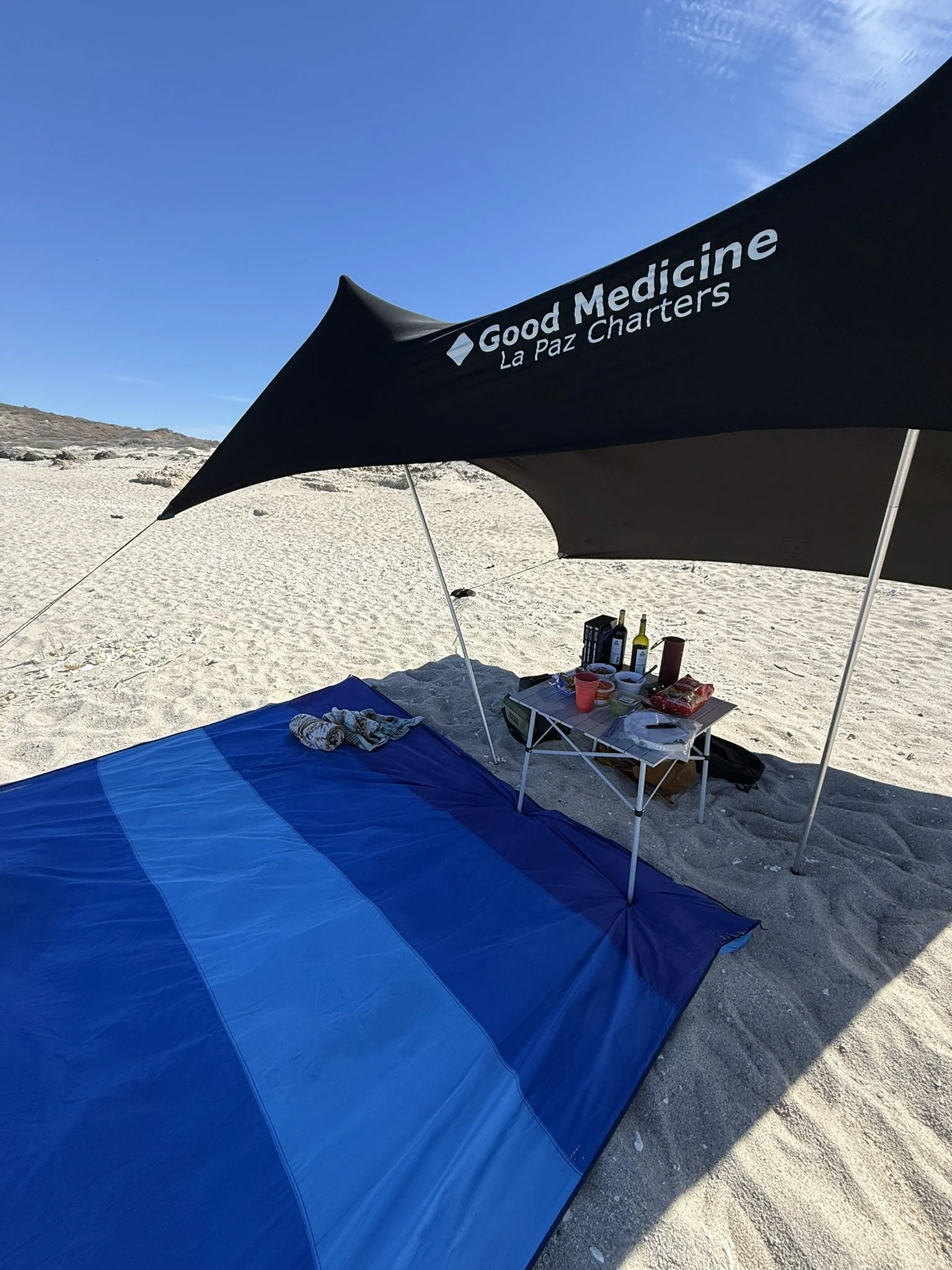 Camping setup on a sandy beach with a large blue sleeping pad, a black canopy with white lettering for 'Good Medicine La Paz Charters,' and a small portable table with bottles, food, and cups.