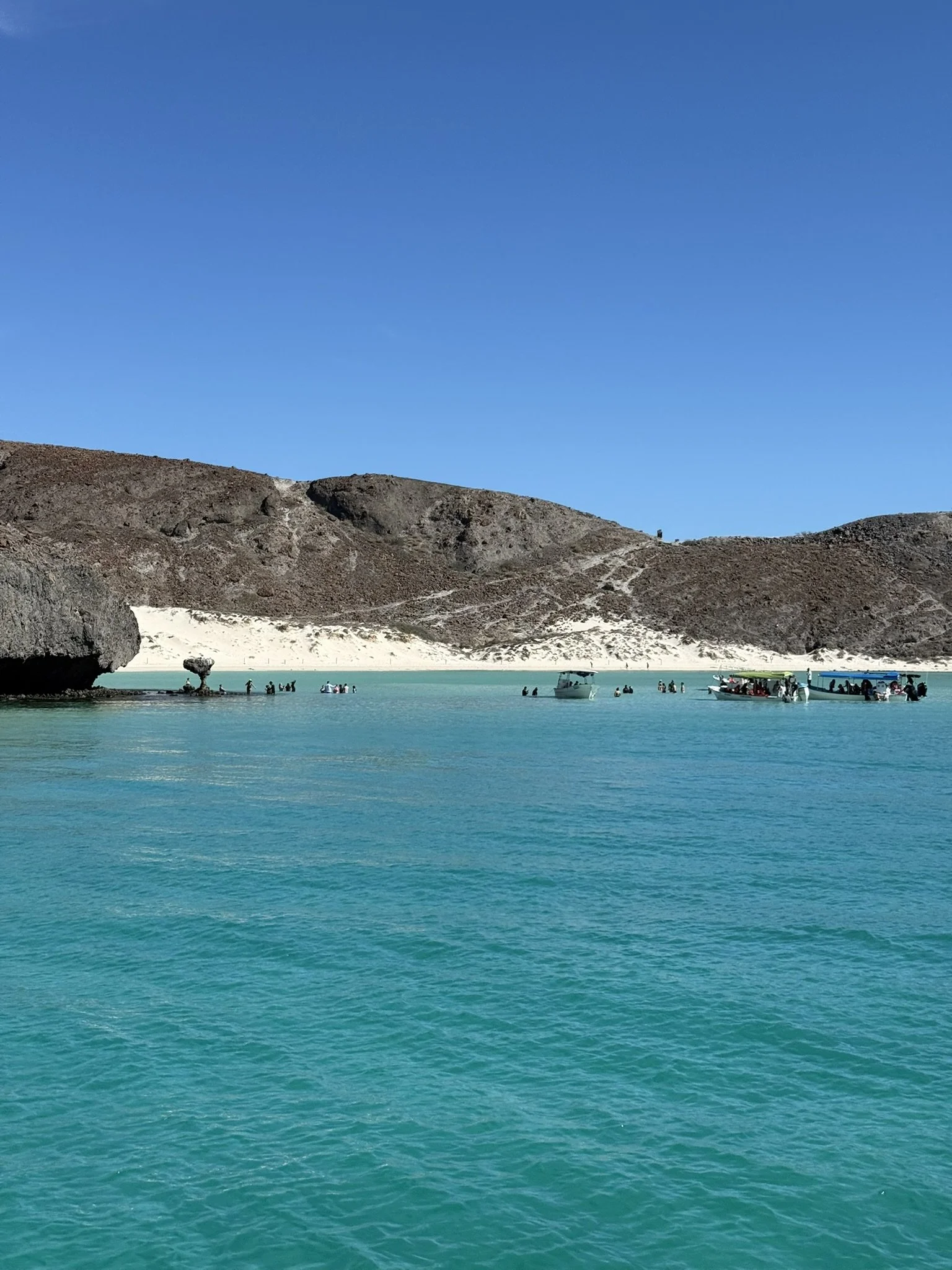A turquoise body of water with a large rock on the left and several boats with people on the water, a sandy hill with sparse vegetation in the background, and a clear blue sky.