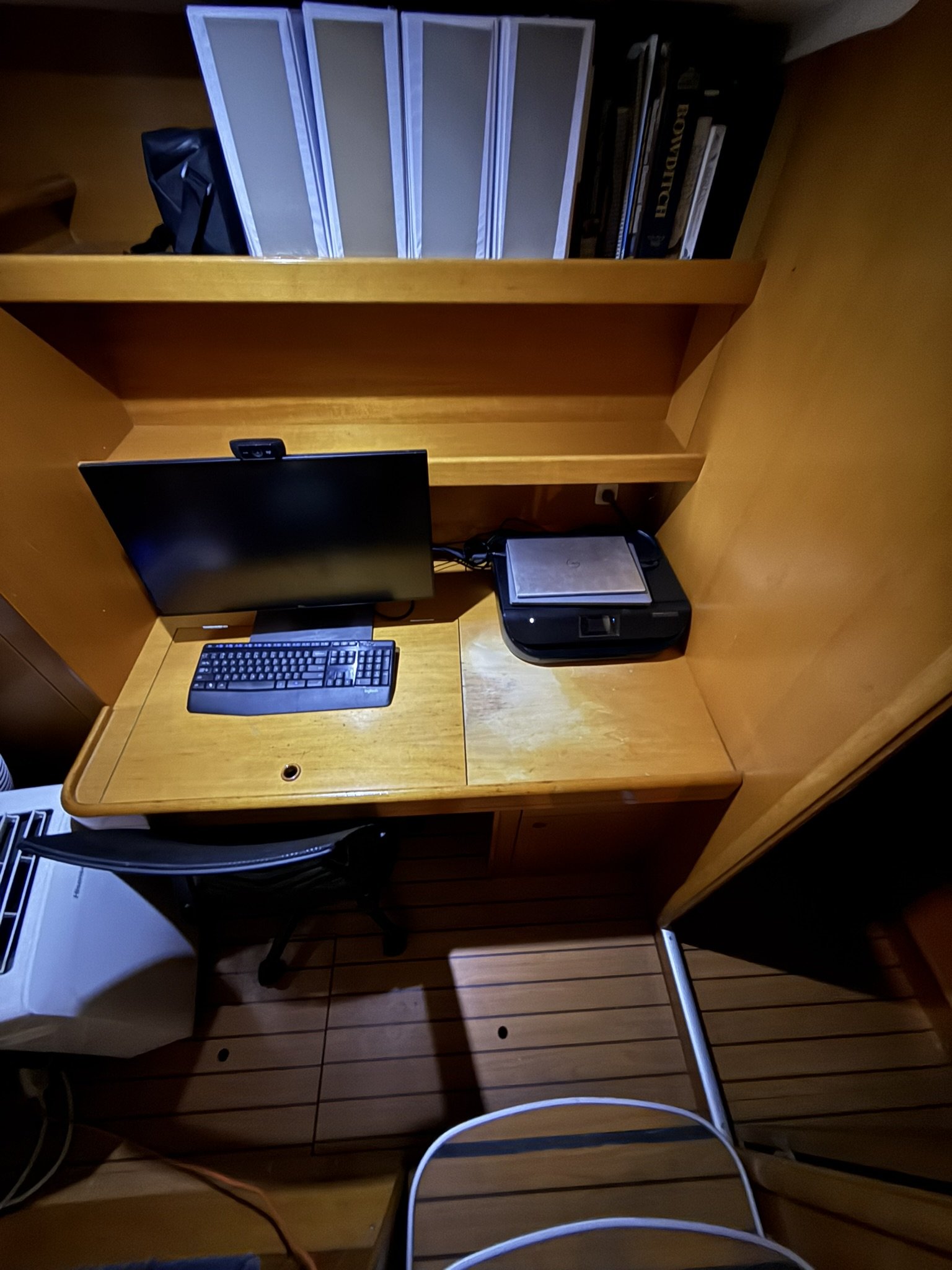 A computer desk with a monitor, keyboard, mouse, and a desktop computer tower. There is a white chair at the desk. Behind the desk, there is a wooden bookshelf with books and some folders.