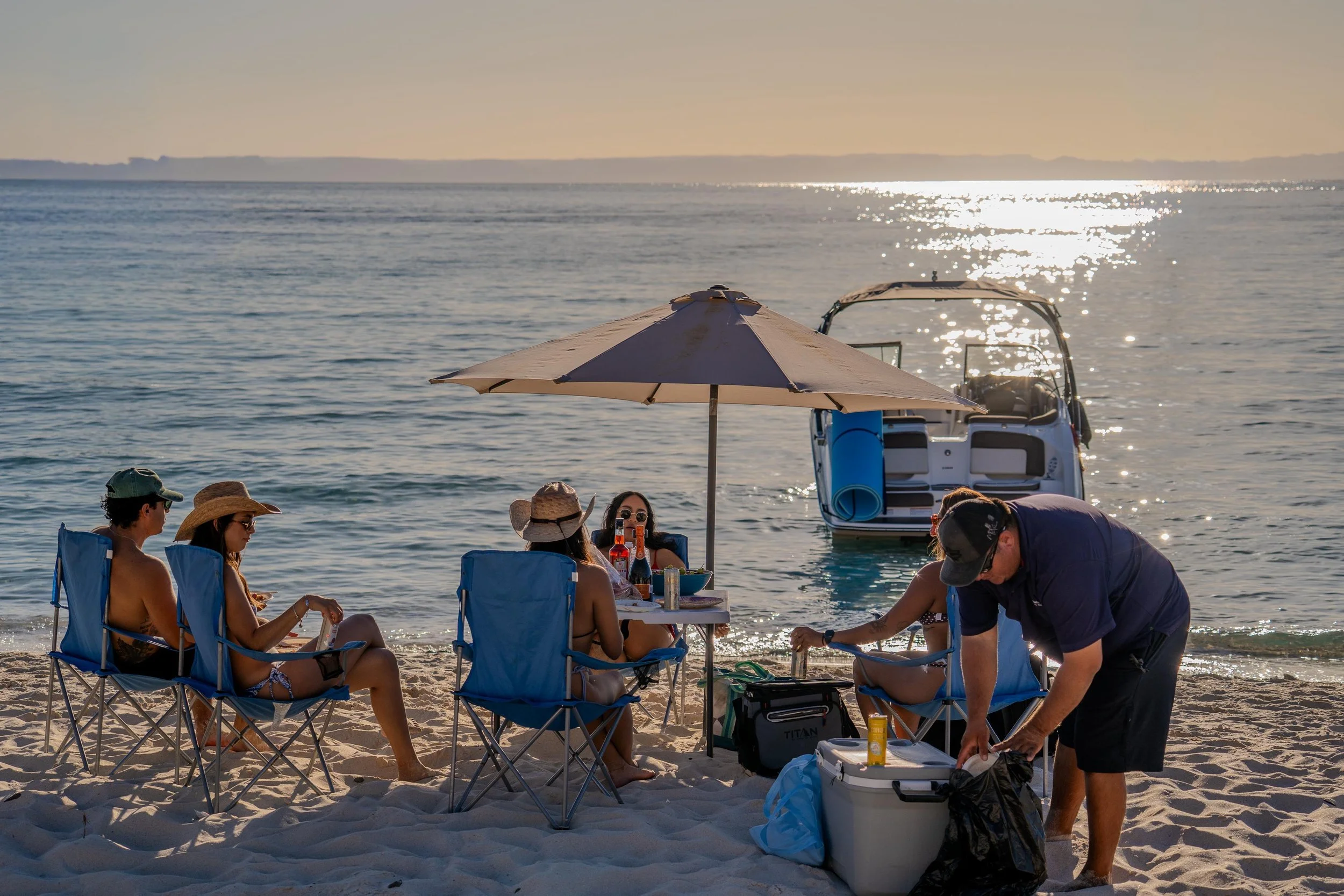 Group of people sitting on beach chairs under an umbrella by the shore, with a boat moored nearby and a sunset reflecting on the water.