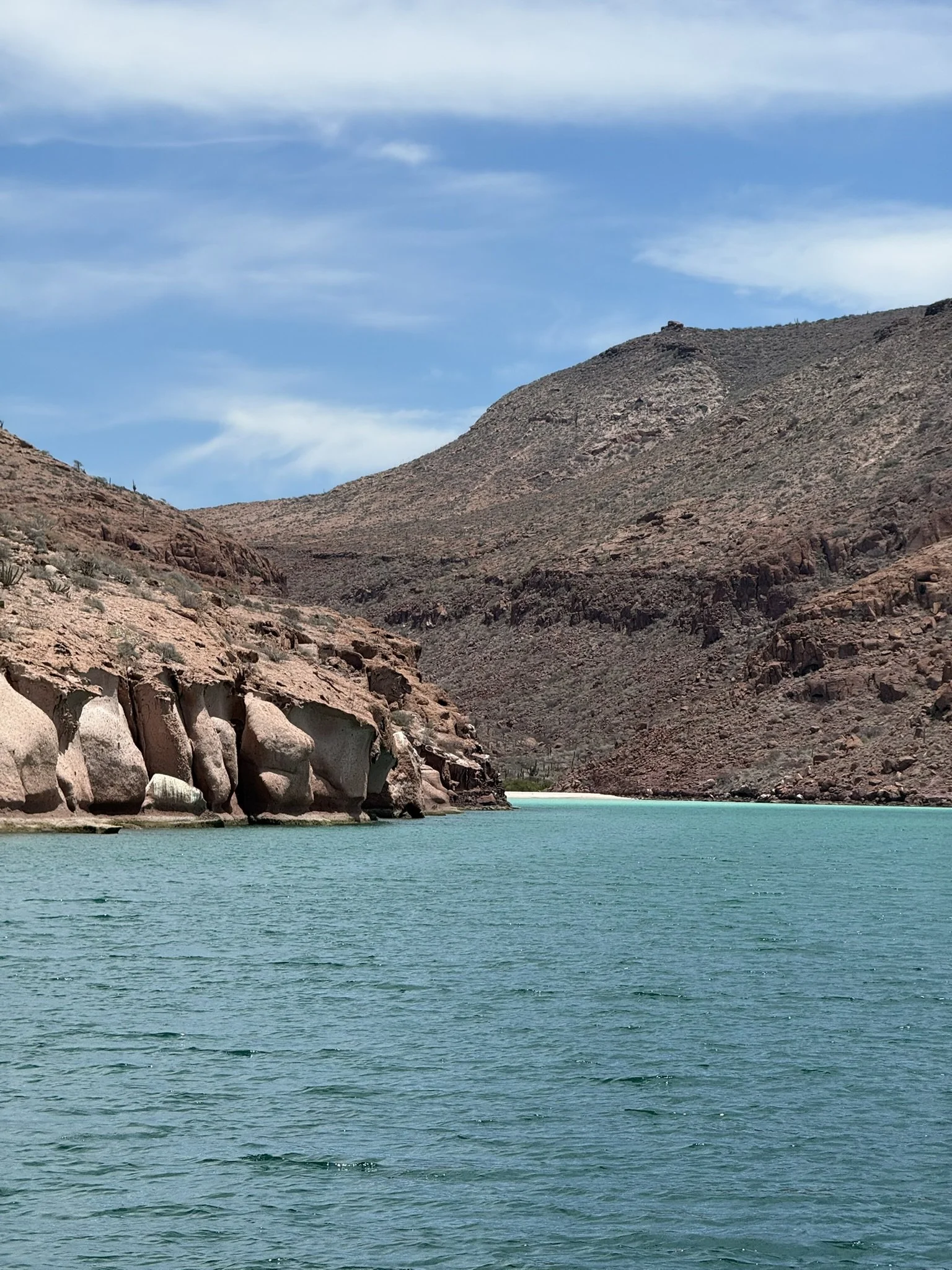 A scenic view of a turquoise body of water bordered by rocky, arid hills with sparse vegetation against a blue sky with scattered clouds.