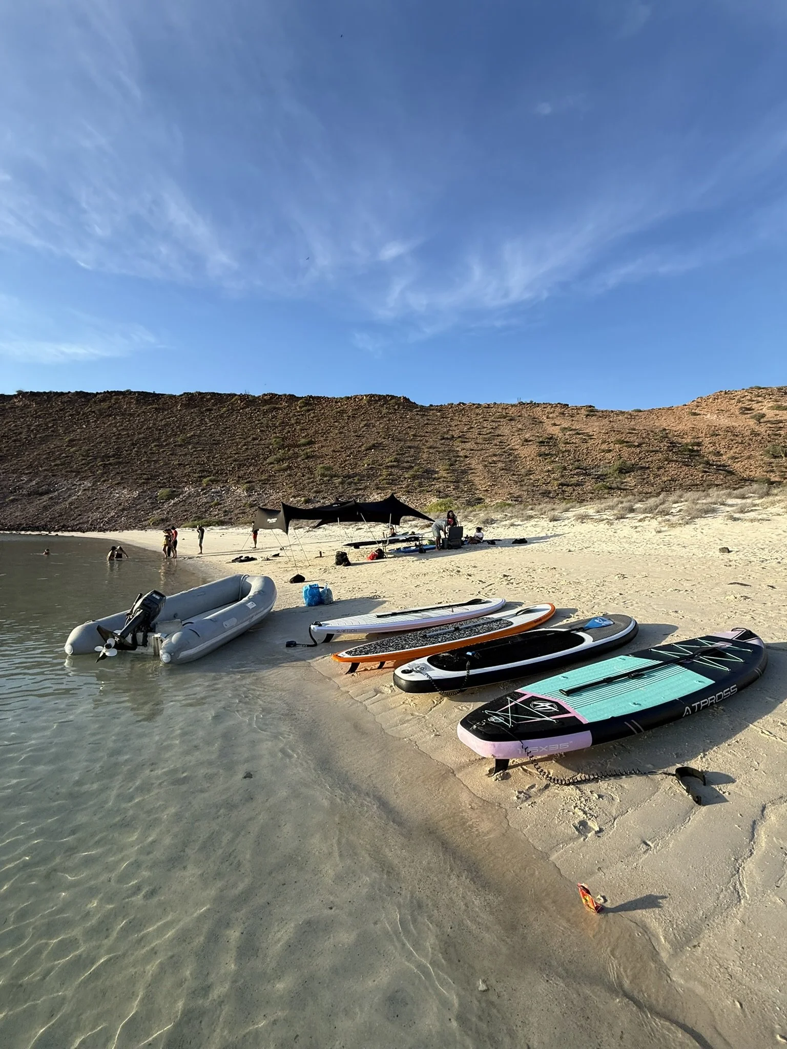 Boats and paddleboards on the sandy shore of a beach with a hill and blue sky in the background.