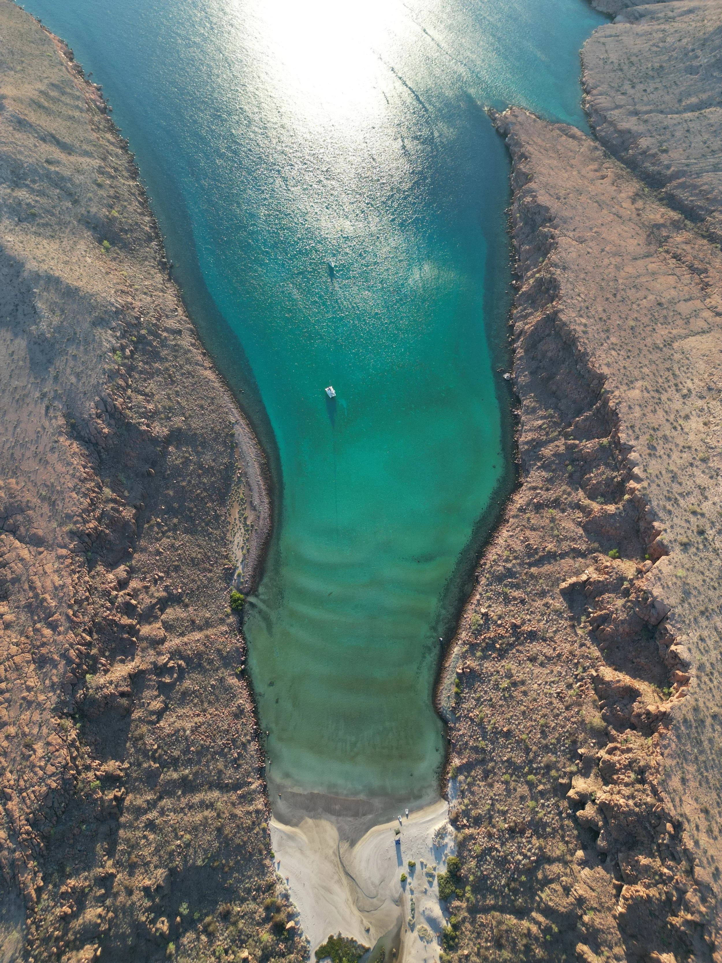 Aerial view of a winding river with clear blue-green water flowing through a dry, rocky canyon, with a boat visible on the water and a small boat ramp at the bottom of the image.