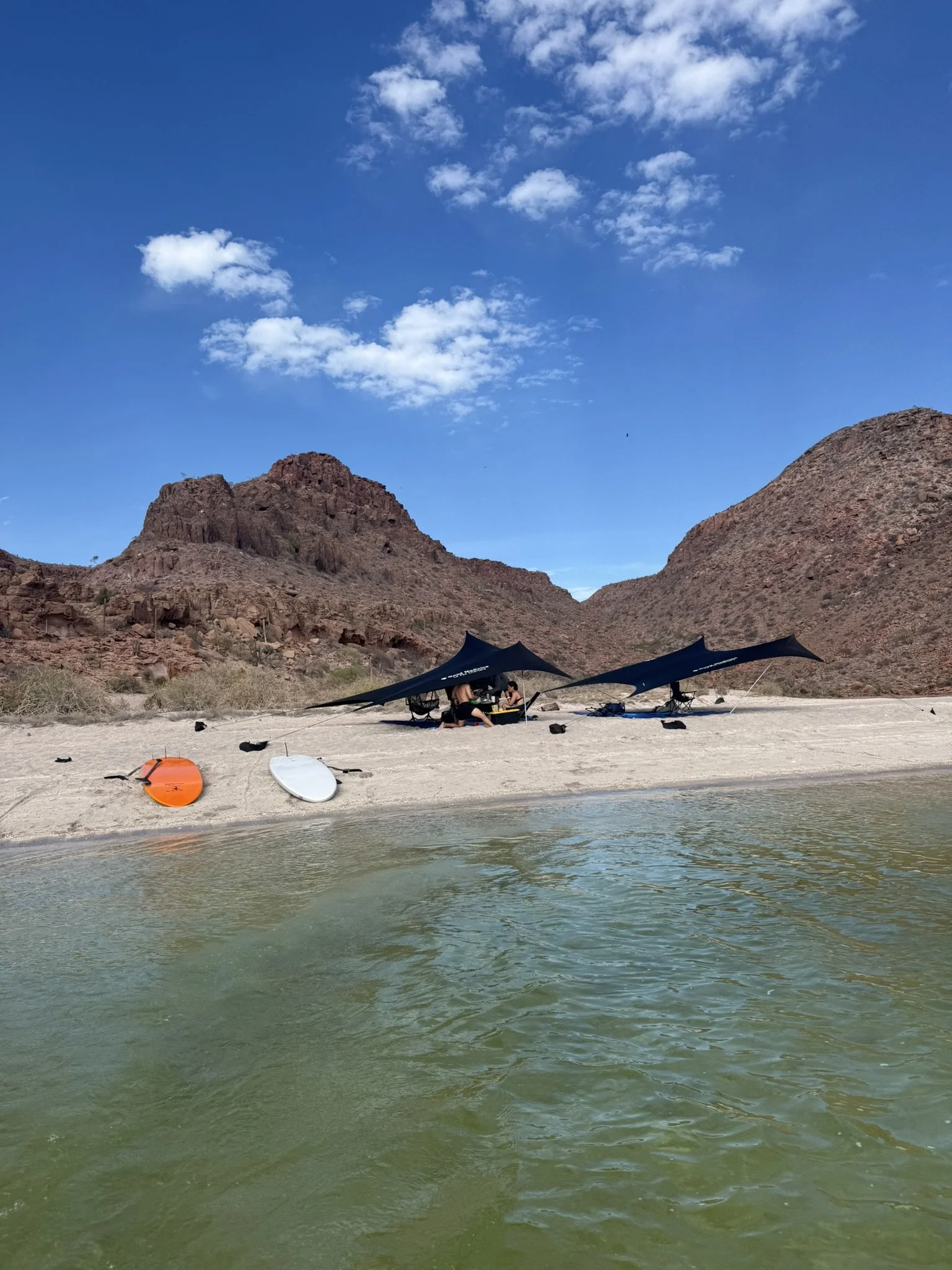 A desert landscape with rocky hills and a blue sky with clouds, featuring a group of people under shade tents near the water, with kayaks and paddleboards on the sandy shore.